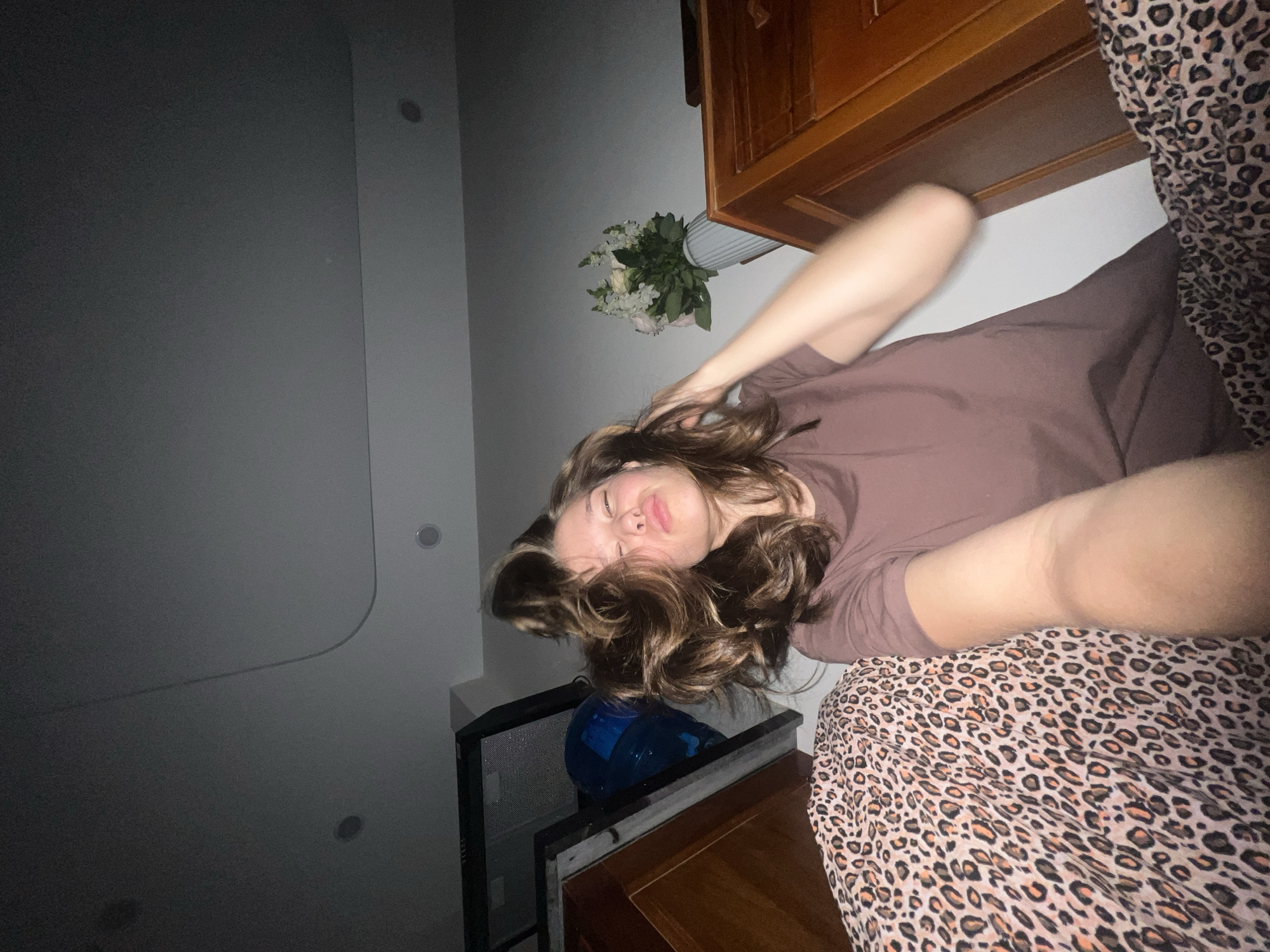 A young woman with brown curly hair and a mauve shirt is lying on a bed with a leopard print blanket. Her eyes are closed. There is a bouquet of white flowers in a vase on the wall behind her. A dark, tall cabinet with wooden drawers is beside the bed. Part of the ceiling is visible, showing recessed lighting. The location is difficult to ascertain from the image but seems to be a domestic indoor setting. There is no indication or evidence related to "Nam Natural Heritage Area Trang, Vietnam."