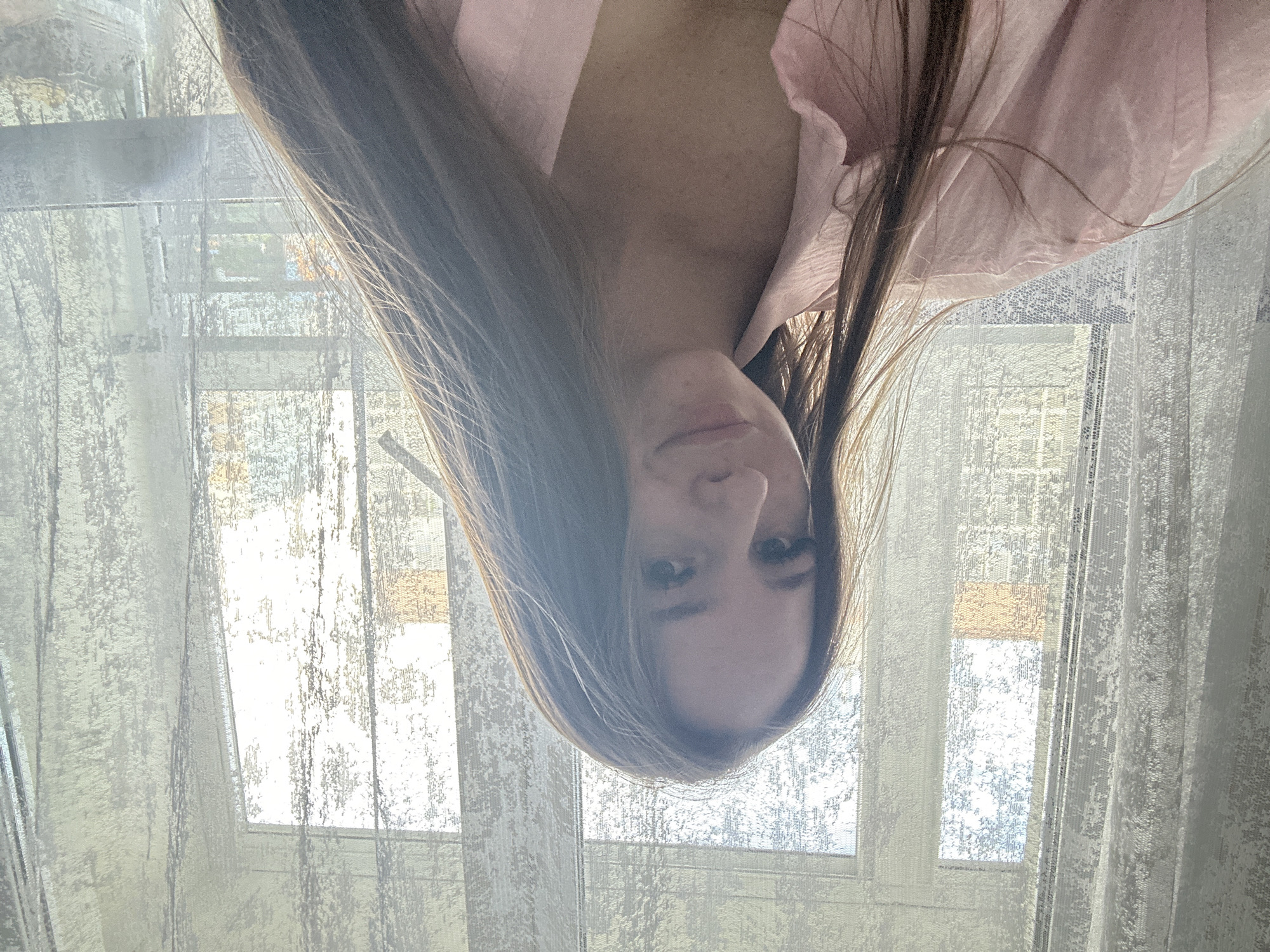The image displays an inverted close-up of a person, likely female, with long, dark brown hair, looking directly at the camera. Their head and upper torso are visible, with hair fanned out around their face. The person is wearing a light-colored, possibly pink or peach, button-up shirt. Their eyes are open, and their expression is neutral, possibly with a faint smile.
In the background, bright natural light streams through windows covered by sheer, light-colored curtains or blinds. The curtains feature a textured or patterned design. The window frames are dimly discernible behind the translucent fabric. The bright illumination suggests it is daytime, possibly a sunny morning or afternoon. The setting appears to be indoors, likely a room with windows.
The activity taking place seems to be a portrait or selfie captured from an unusual, inverted perspective. There are no specific location cues visible, apart from the indoor setting. No discernible text is present in the image.