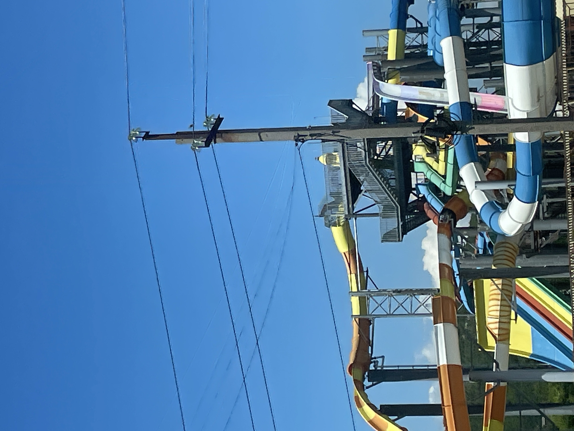 The image shows a large water park with multiple slides of various colors and shapes. The slides are made of plastic and metal, and they are arranged in a complex structure with walkways and stairs. The water park is located outdoors, and the sky is blue with some clouds. The image also shows a utility pole with power lines in the foreground. The water park is likely a popular tourist attraction in Lermontovo, Russia.
