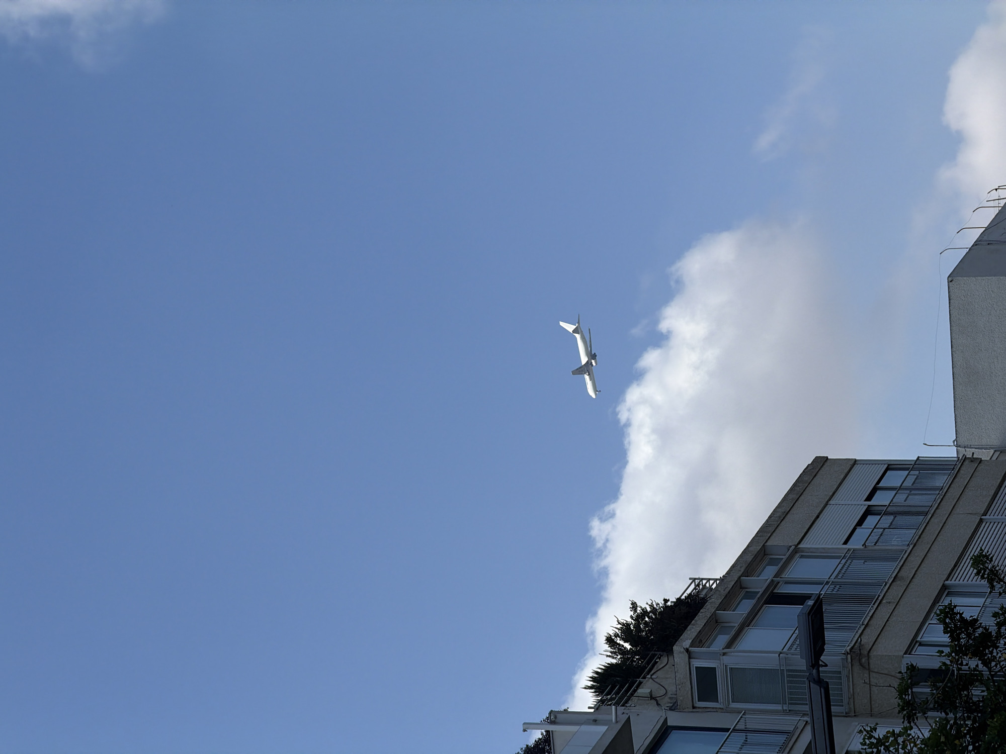 The image depicts a clear blue sky with scattered white clouds. In the upper right quadrant, a white airplane is visible in flight. In the lower left portion of the image, the corner of a modern building complex is shown, characterized by concrete structures, glass windows, and balconies with metal railings. Trees and greenery are present on the rooftop of the building. The scene appears to be an urban environment, likely in Tel Aviv, Israel, given the architectural style and clear sky often observed in the region. There are no people visible in the image. The overall impression is one of a bright, sunny day in a city.