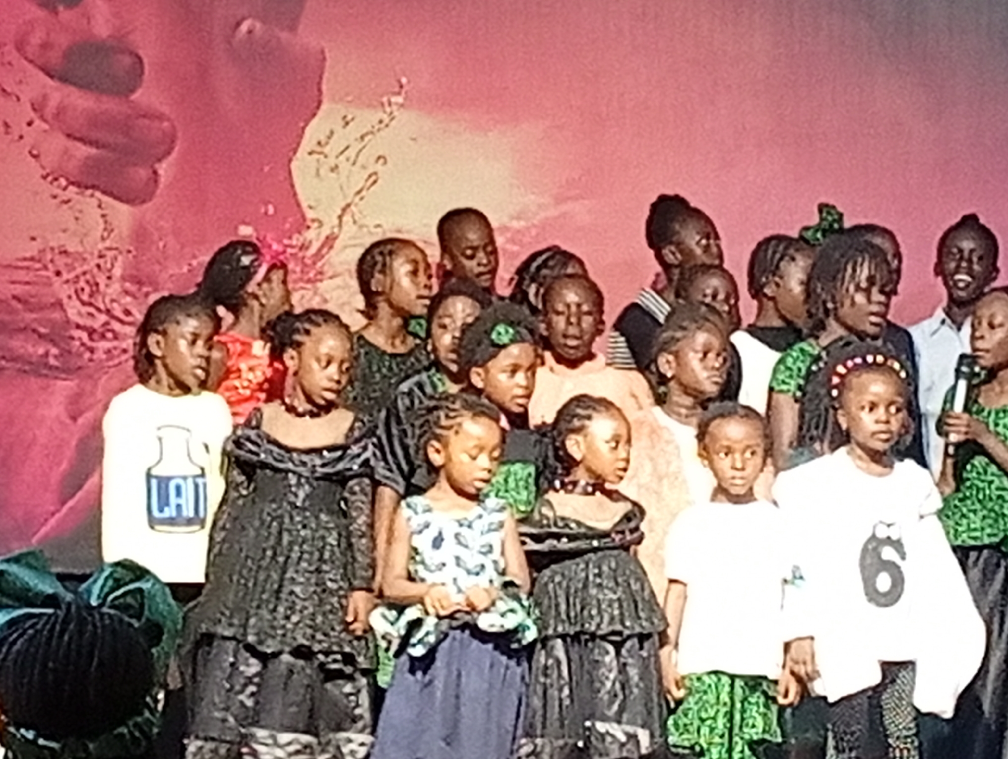 A group of approximately twenty young Black children, appearing to be between the ages of five and twelve, are gathered on a stage. They are standing close together, facing the viewer, and seem to be participating in a performance or presentation. The children are dressed in a variety of outfits, including traditional African attire, dresses, and t-shirts. Some are wearing black, lacy dresses, while others have on colorful patterned clothing. One child in the front row is wearing a white t-shirt with a large black number '6' printed on it. Another child to their left is wearing a white t-shirt with a graphic of a milk bottle and the word "LAIT" in blue. The background is a soft pinkish-orange, with a blurred image of a hand and some text. The lighting is bright and appears to be from stage lights. The overall impression is of a lively cultural event, possibly a school play or community gathering. The image was taken at the National Museum, Jos, Nigeria.