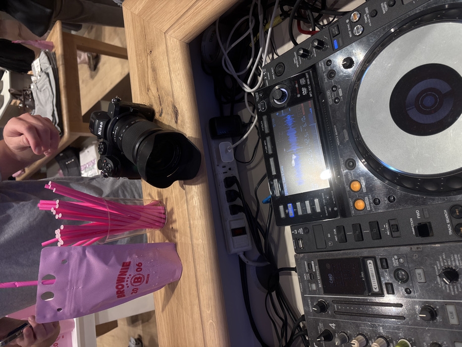 The image displays an indoor event setup in the city of Unknown, Unknown. On a light wooden counter, a pink beverage pouch with a pink straw, branded "BROWNIE 20 B 06 MEXICO", stands to the left of a clear glass filled with numerous pink straws. To the right, a black Nikon mirrorless camera with a zoom lens and lens hood rests on the counter. The hands and upper torso of a person in a grey shirt are partially visible behind the drink and straws. The blurred background shows other individuals and a wooden table with folded garments, suggesting a retail or event space. Directly beneath the counter, a white power strip is connected to several plugs and a tangle of black cables. Below this, a DJ setup is active, featuring a mixer unit on the left and a media player unit on the right. The media player's screen displays a blue audio waveform, indicating "DJ RELAX: Take It Easy", along with time and other interface elements.