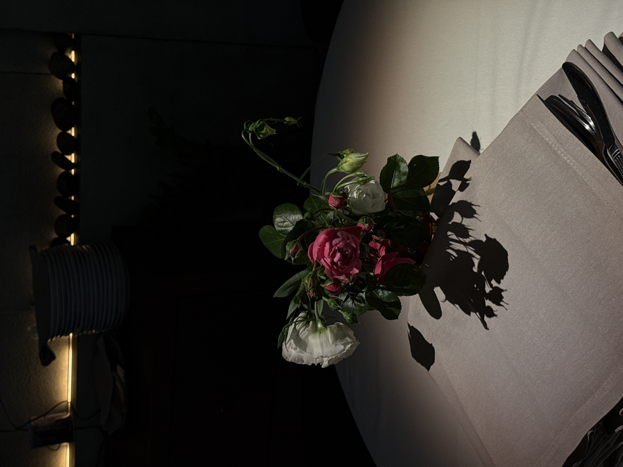 The image depicts an intimate dining setting, likely in a restaurant or a sophisticated home, characterized by dramatic, low lighting. The central subject is a small bouquet of fresh flowers, predominantly pink roses, white roses, and other white blooms, with vibrant green foliage. These flowers are placed on a light-colored, possibly light gray or beige, tablecloth or placemat.
A strong, directional light source illuminates the flowers and a portion of the table, casting prominent, dark shadows of the bouquet onto the tablecloth, adding depth and a stylized feel to the composition. To the right of the flowers, a folded napkin holds a set of polished silverware, including a fork and a knife. The table itself appears to be round or oval, with a curved edge visible in the background, also illuminated by the same light source.
On the far left, in the darker background, a vertical strip of warm, golden light illuminates a series of dark, circular decorative elements, creating a soft glow. Below this, a stack of white plates is partially visible, suggesting a ready setting for a meal. The overall ambiance is moody and elegant, indicative of an evening setting. No people are visible in the frame, and there is no discernible text. The scene evokes a sense of quiet luxury and prepares for a special occasion.