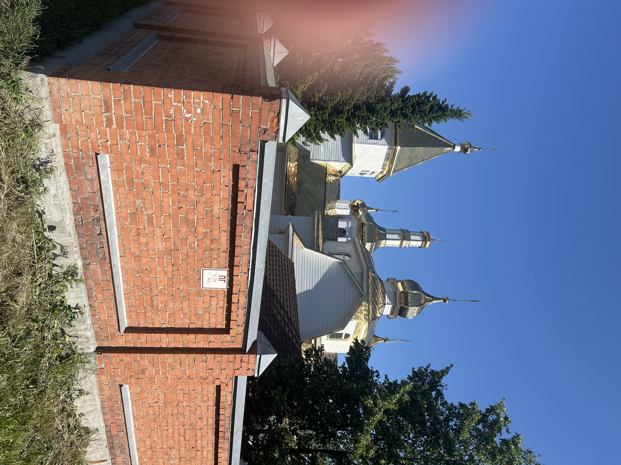 The image depicts a bright, sunny day with a clear blue sky. In the foreground, a red brick wall with concrete foundations and a metal coping runs horizontally across the frame. A small white sign with Cyrillic lettering and a symbol is affixed to the wall. Behind the wall, a white building with golden domes, characteristic of a church, is visible. Several tall green coniferous trees and a large leafy deciduous tree surround the building, partially obscuring it from view. The building appears to have a traditional architectural style with multiple domes and spires. The overall impression is that of a religious building in a semi-urban or suburban setting. The image is dated July 11, 2023. The church in the image is the Church of St. Nicholas the Wonderworker, located at Lenina Street, 54, Kemerovo, Russia.