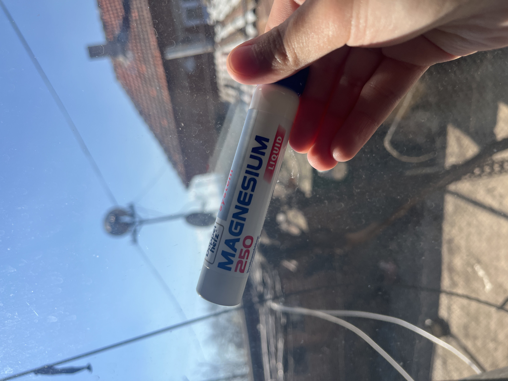 A hand holds a white tube labeled "Magnesium Liquid 250" against a window. Through the window, there is a view of a partially visible house with red roof tiles and blue sky. Also visible through the window is what seems to be an object on a pole. The location cannot be determined from the image.