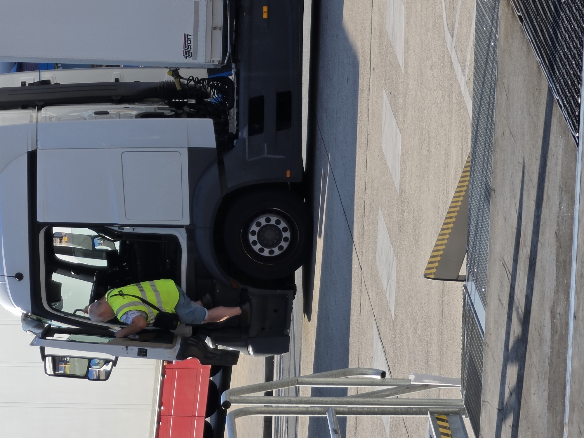 A middle-aged man wearing a bright yellow safety vest and blue shorts is climbing into the cab of a white semi-trailer truck. The truck has a black bumper and black side panels. The man is using the steps to enter the cab. The truck is parked on an asphalt surface, possibly at a logistics center or industrial area. In the background, another white vehicle, possibly a trailer, is visible. The image appears to be taken during the daytime, with bright sunlight casting shadows on the ground.