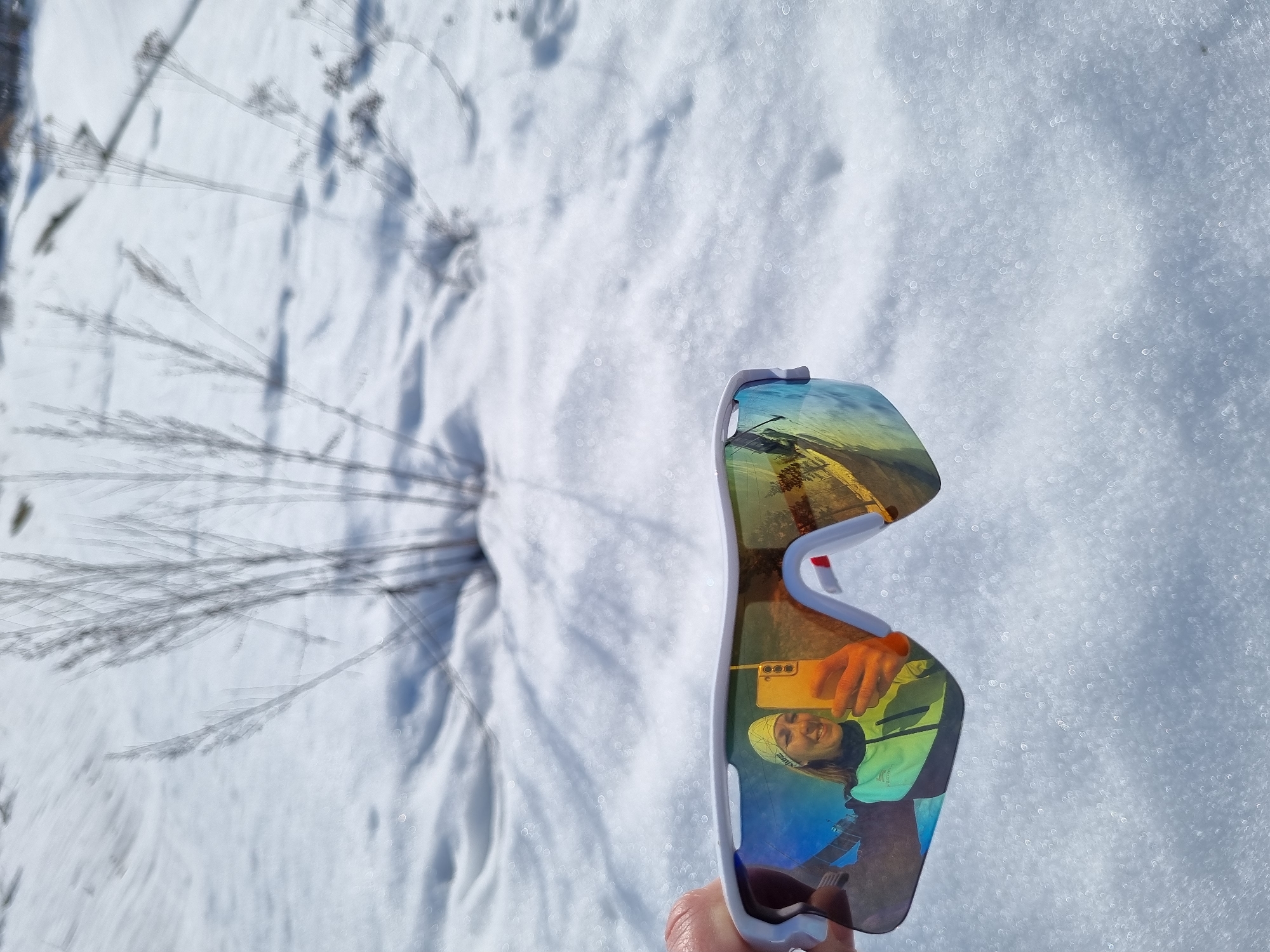 The scene depicts an outdoor, snow-covered environment in Chaikovsky, Russia, under bright daylight conditions. In the lower foreground, a hand holds a pair of white-framed sports sunglasses featuring iridescent, mirrored lenses that reflect blue, green, yellow, and orange tones. The left lens reflects a smiling woman wearing a yellow beanie and a light green and black athletic jacket. This reflected woman holds a light-colored smartphone, visible with its camera array, pointed towards herself. The right lens also reflects a portion of the smartphone and a blurred outdoor background, including what appear to be distant trees or structures. The midground and background consist of a field of white snow with subtle undulations. Several clusters of dried, light-colored vegetation stalks protrude from the snow, casting distinct shadows onto the surface, indicating direct sunlight. These background elements are out of focus.