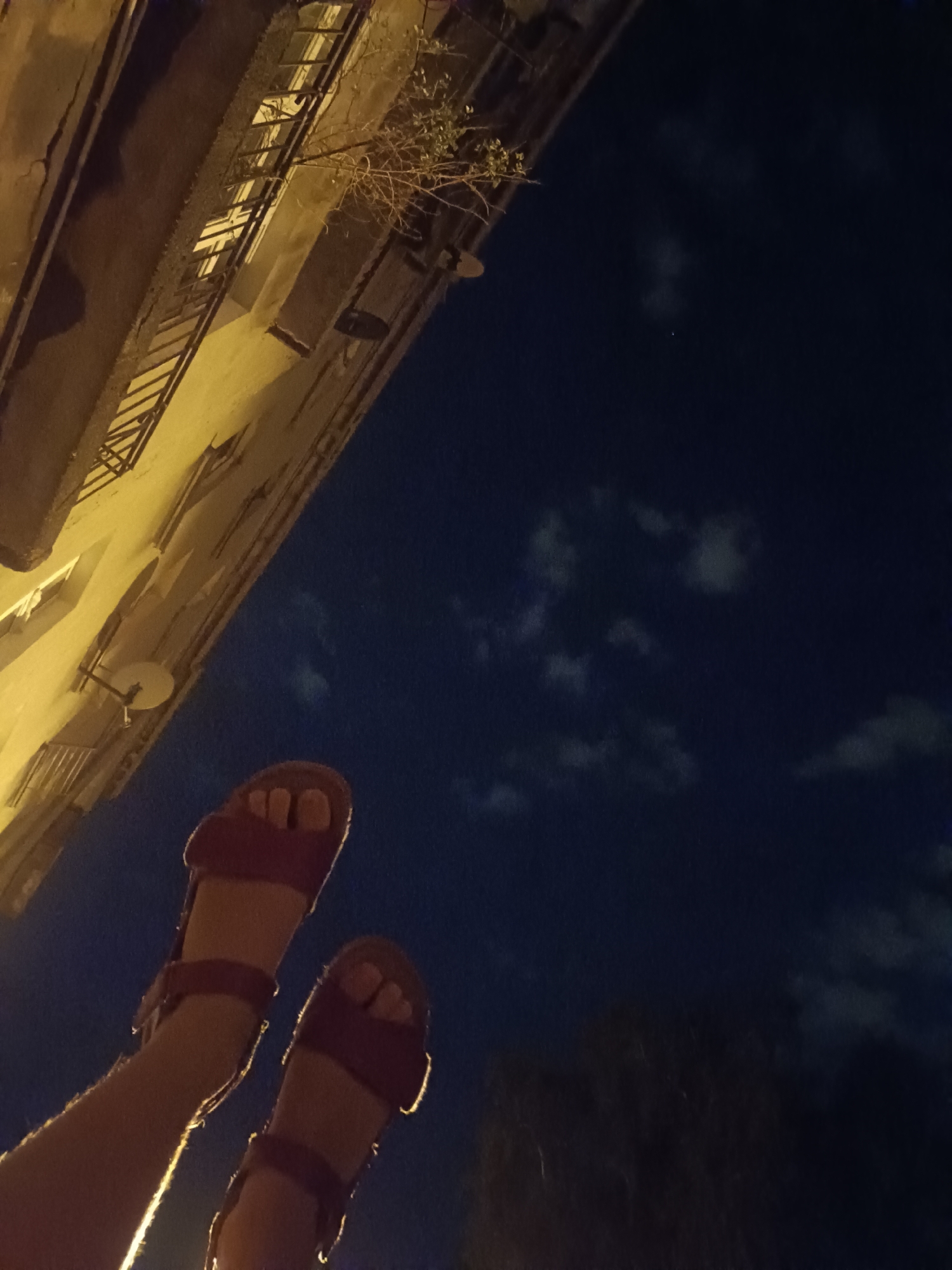 The image depicts a low-angle, night-time view of two bare feet wearing dark red sandals, positioned in the foreground and extending upwards. The feet are illuminated by a warm, yellowish light, highlighting the texture of the skin and the sandals. Behind the feet, a building facade is visible, characterized by its weathered, pale yellow plaster and dark metal railings along the windows. A sparse, dry branch with a few green leaves extends from the facade, partially obscuring a window. The background is a deep blue night sky, with scattered, wispy clouds illuminated by an unseen light source. A small, faint point of light, possibly a star or distant aircraft, is visible in the sky. The scene appears to be set in an urban environment, potentially a residential street or courtyard, typical of cities like Łódź, Poland, known for its historic architecture. There are no other people or discernible interactions visible in the image. The overall atmosphere is still and quiet, conveying a sense of late evening or night.
