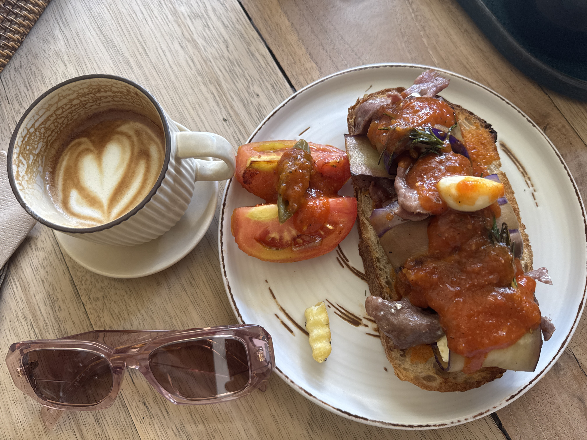 The image depicts a table at a cafe in an unidentified location. A cup of coffee with latte art, resembling a heart, rests on a saucer beside a white plate. On the plate is an open-faced sandwich topped with what appears to be slices of meat, possibly beef or lamb, vegetables, and a red sauce. Sliced tomatoes accompany the sandwich. A single, golden-brown French fry sits separately on the plate. To the left of the coffee cup, a pair of pink-framed sunglasses lies on the table. The table is made of wood, with visible grain.