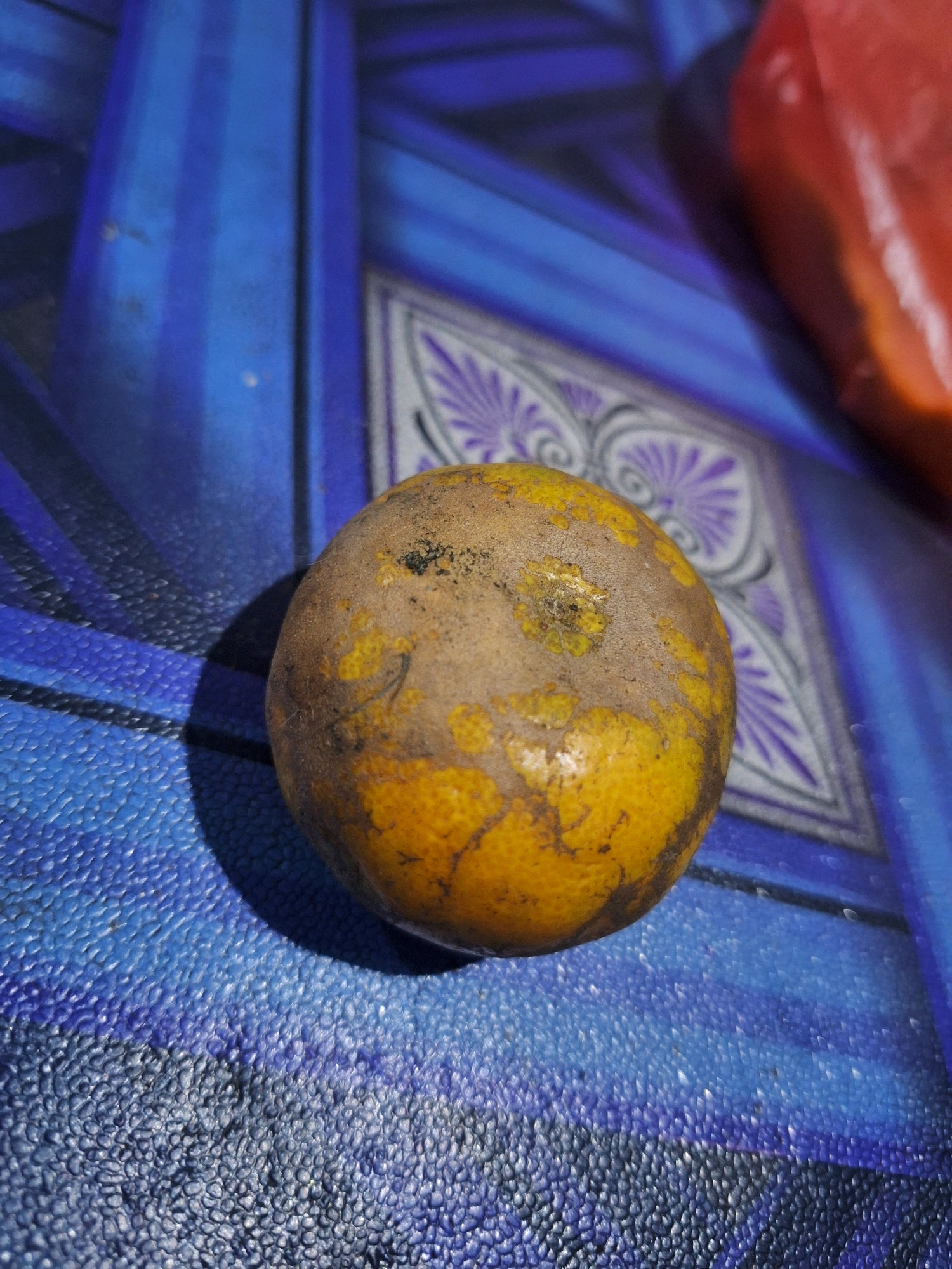 A single, unpeeled orange sits on a blue, textured surface with a patterned background. The orange has a predominantly yellow peel with areas of brown and some rough texture, suggesting it might be slightly aged or have some imperfections. The blue surface appears to be a placemat or tablecloth with a pattern that includes vertical stripes and some decorative, possibly floral, elements in purple and gray. In the upper right corner of the image, a blurred red object is visible, which could be another item of food or a bag. The lighting casts a shadow to the left of the orange. The overall setting is a still life arrangement, with no people or specific landmarks visible to pinpoint a location within Air Tawar, Indonesia.