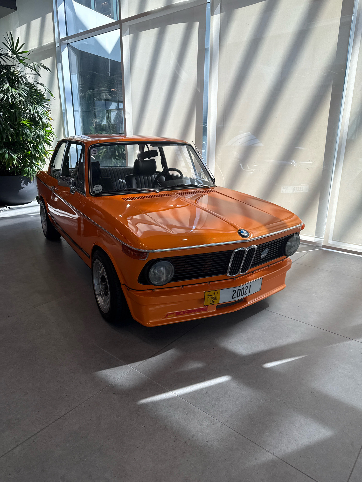 An orange 1973 BMW 2002 is parked on a gray tiled floor in an indoor location in Dubai, United Arab Emirates. The car has a black interior and a black license plate frame. The license plate reads "20021" and has a small yellow plate with "DUBAI CLASSIC" on it. The car is positioned facing slightly to the left, with its front end angled towards the viewer. Behind the car, a large window is covered by a beige blind with diagonal light patterns cast upon it. To the left of the car, a large potted plant with green leaves is visible. The environment appears to be a modern building with large windows and a clean, minimalist design.