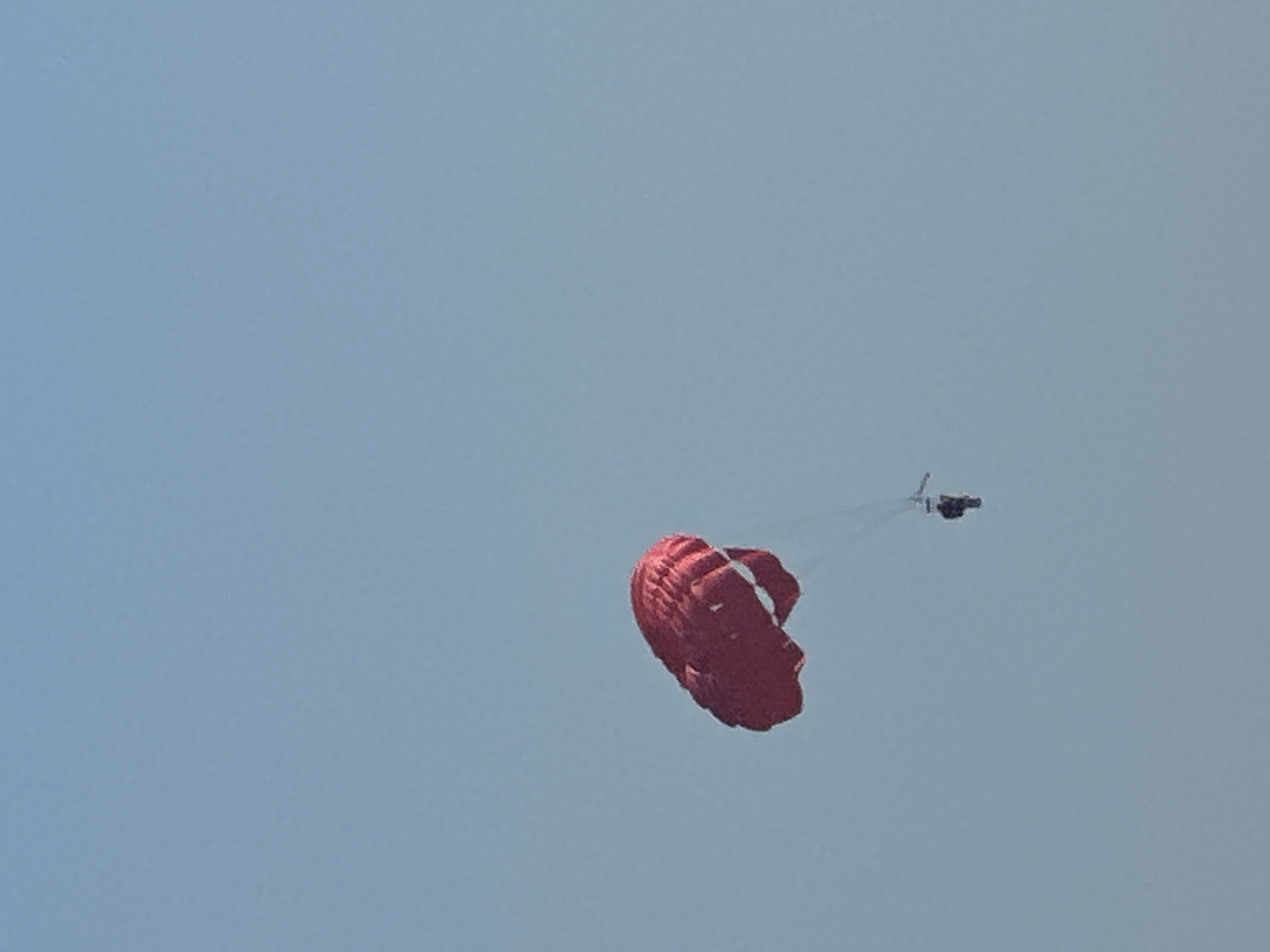 A person is parasailing in the sky above the city of Boreti, Montenegro. The parasail is red and appears to be filled with air. The person is suspended below the parasail by ropes. The sky is clear and blue.