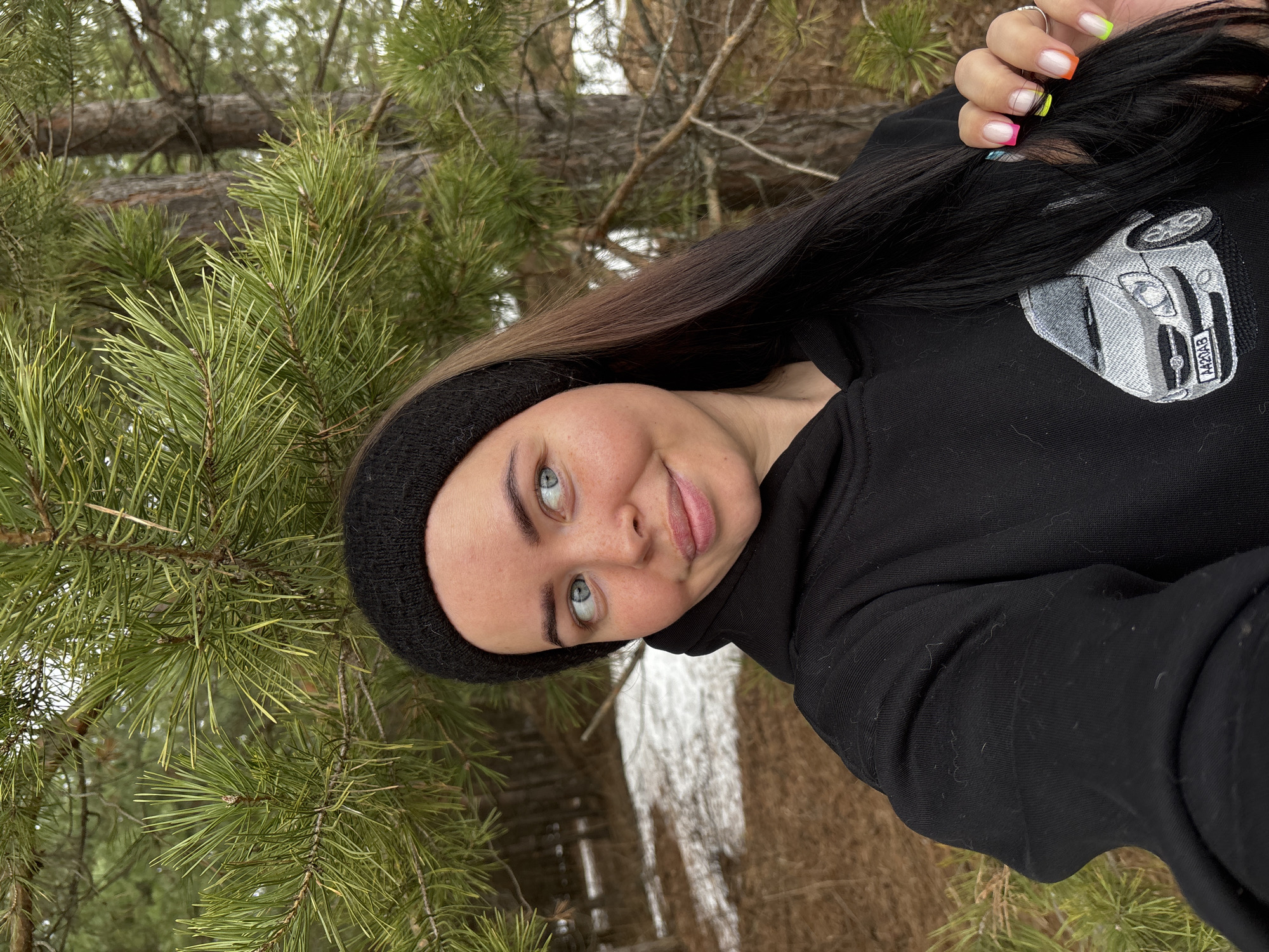 A person is in a pine forest in Русский Вожой, Russia. They are wearing a black hoodie with an embroidered car on it and a black headband. They have long hair that is dark at the roots and lighter at the ends. The person's nails are painted in bright colors. The person is surrounded by pine trees and the ground is covered in snow.