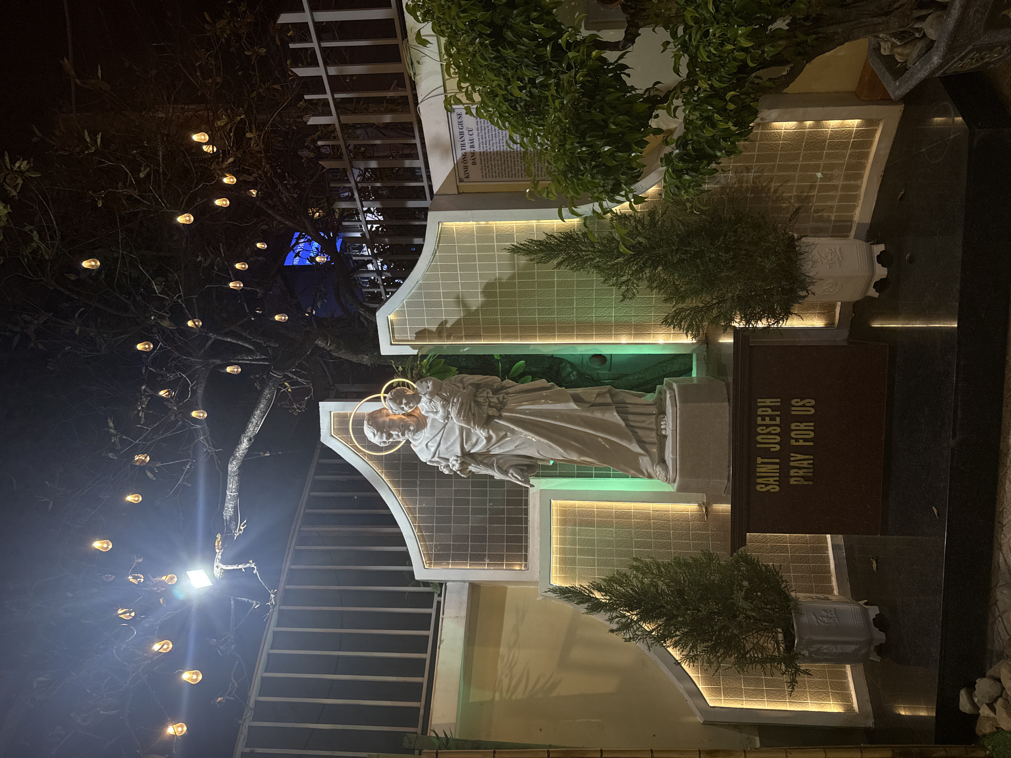 The image depicts a statue of Saint Joseph holding the infant Jesus, illuminated by LED lighting at night. The statue stands in front of a wall with a grid-like pattern, bordered by more LED lighting. Green plants are positioned on either side of the statue. A dark brown sign to the right of the statue reads "SAINT JOSEPH PRAY FOR US." In the background, a tree with hanging light bulbs is visible to the left. A metal staircase is also visible in the background to the left of the statue. Based on the statue of Saint Joseph and the prayer, this is likely a religious site. According to information associated with the image, it is located in Nha Trang Ward, Vietnam.