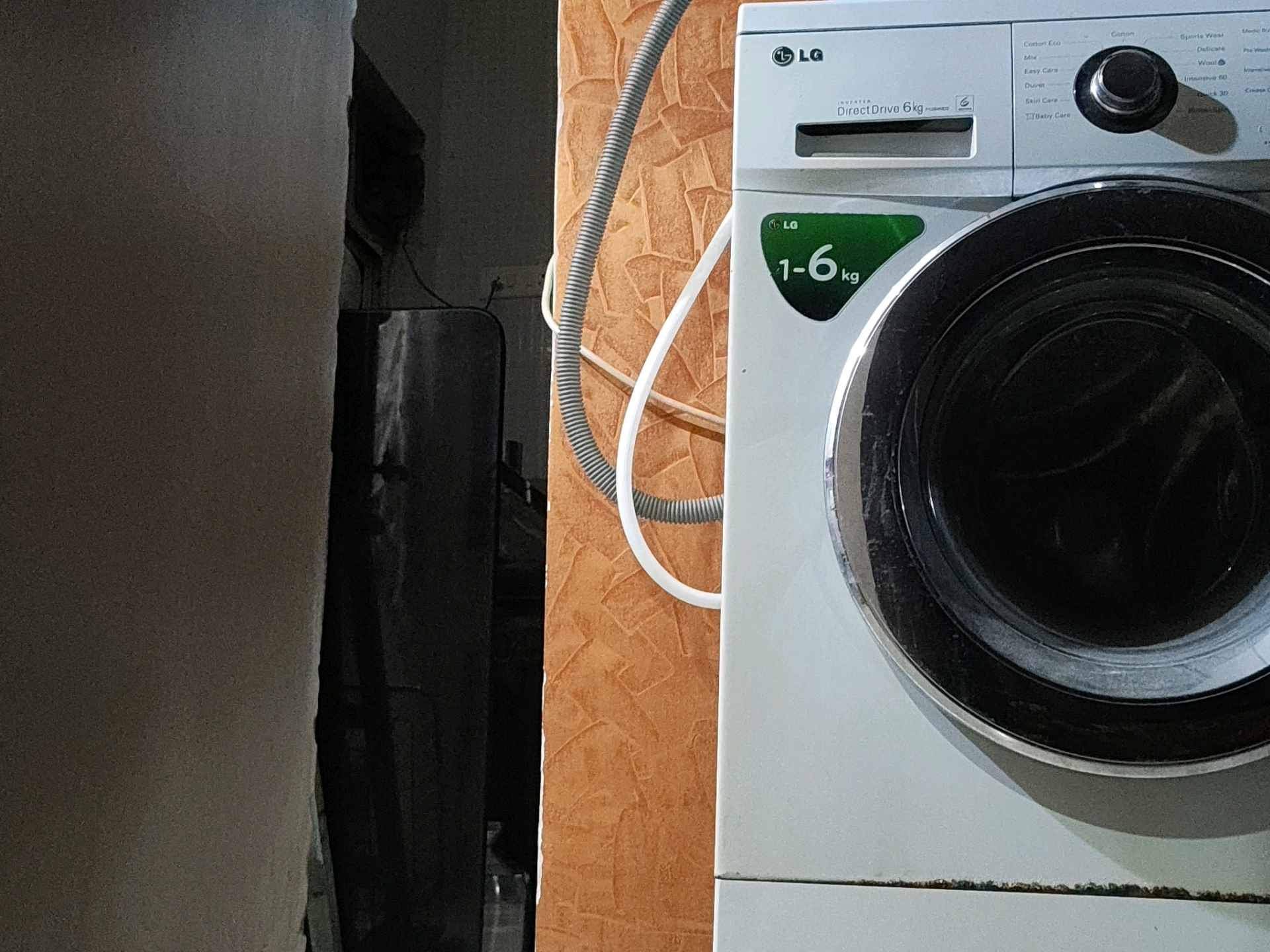 In Pune, India, this image captures a close-up view of a white LG washing machine. The washing machine is positioned in a room with textured orange walls. A gray corrugated hose and a white flexible hose are visible, connected to the washing machine. To the left of the washing machine, a dark, tall object, possibly a laundry hamper or cabinet, stands against a neutral-colored wall. The lighting suggests an indoor setting, likely a laundry area or kitchen. The focus is on the washing machine's control panel, which displays various wash cycle options like "Cotton Eco," "Mix," "Easy Care," and "Baby Care." The "1-6kg" capacity indicator is also prominently displayed on a green label. The scene is static, with no people or apparent activity.