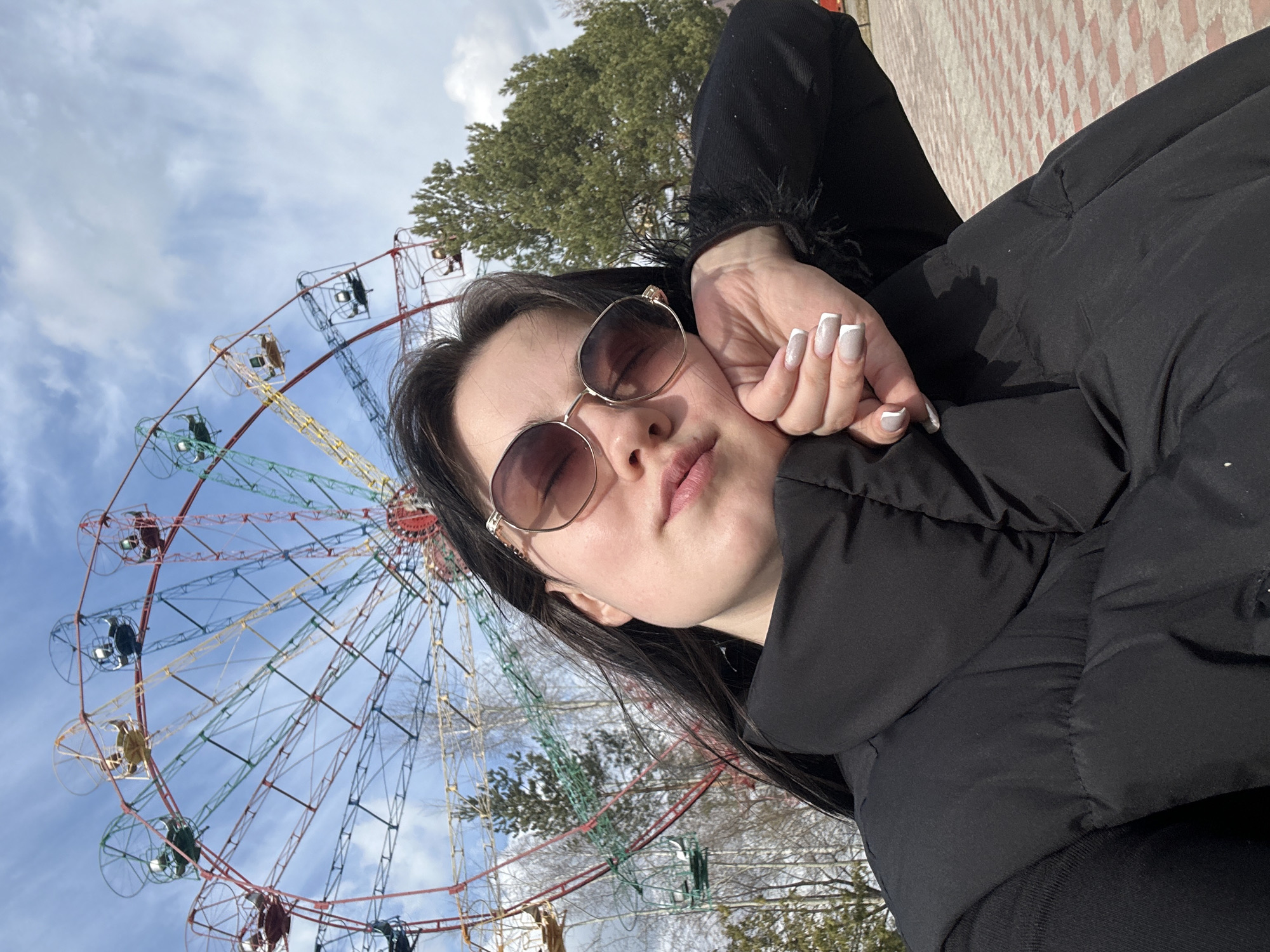 A rotated selfie captures a young woman outdoors in Chaikovsky, Russia, against the backdrop of a large Ferris wheel. The woman, shown from the chest up, has long dark hair and wears dark, round sunglasses and a black puffer jacket. She poses with her head tilted, lips slightly puckered, and her left hand gently resting near her face, revealing nails with a white French tip design.
In the background, a colorful Ferris wheel dominates the left side of the frame, featuring a metallic structure with spokes and gondolas in shades of red, blue, green, and yellow. The sky is bright blue with scattered white clouds, suggesting daytime. Trees with both green foliage and bare branches are visible behind and around the Ferris wheel. A patterned, reddish-brown surface, possibly a brick wall or paved area, is seen in the upper right. The scene appears to be taken in an amusement park or outdoor recreational area. No discernible text is visible in the image.