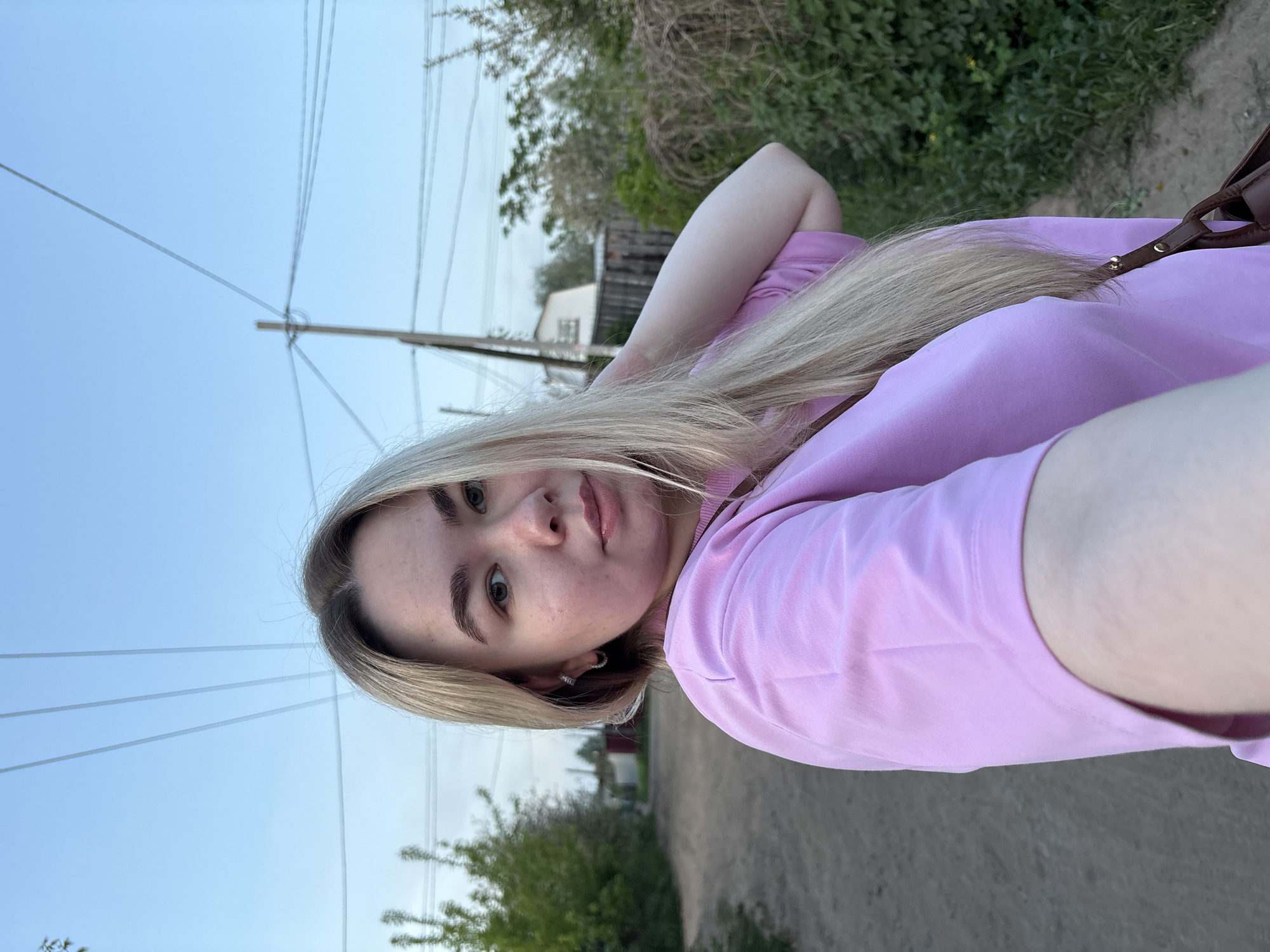 A young woman is taking a selfie outdoors. She has light blonde hair, fair skin, and is wearing a light pink long-sleeved shirt. She is standing on a dirt road in an area with vegetation on either side, appearing to be a residential or semi-rural part of the city of Chaikovsky, Russia, given the presence of power lines and houses in the background. The sky is visible in the upper left corner, suggesting it is daytime. The overall setting suggests a casual outdoor scene.