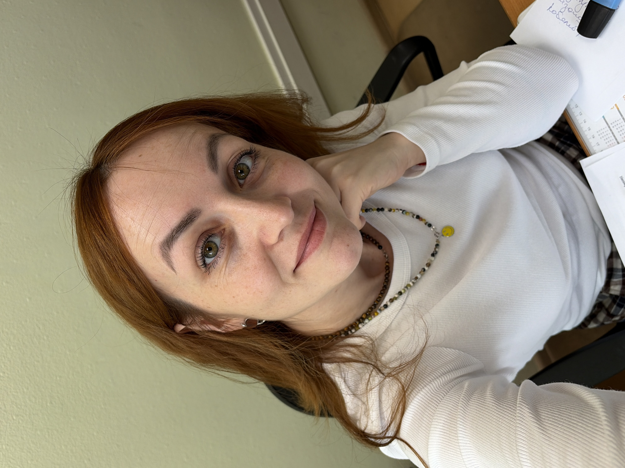 A high-angle, medium-close-up selfie depicts a young woman with long, wavy reddish-brown hair and bright green eyes, looking directly at the camera with a gentle, engaging smile. She has fair skin with visible freckles and wears a white, ribbed long-sleeved top. Around her neck is a multi-colored beaded necklace, featuring tones of brown, black, white, yellow, and green, with a distinct yellow smiley face charm. A small silver earring is visible on her left ear. Her left hand is casually resting near her chin.
The background and setting suggest an indoor environment, likely an office or study in Chaikovsky, Russia. A light, muted green wall is behind her, and a part of a black office chair is visible. To her right, on a wooden desk surface, are several sheets of paper. Some papers contain handwritten text in Cyrillic script, with words like "рабочих" (meaning "working" or "workers") identifiable. A blue and black highlighter and a section of a calendar or planner are also on the desk. The lighting is soft and consistent with an indoor setting.