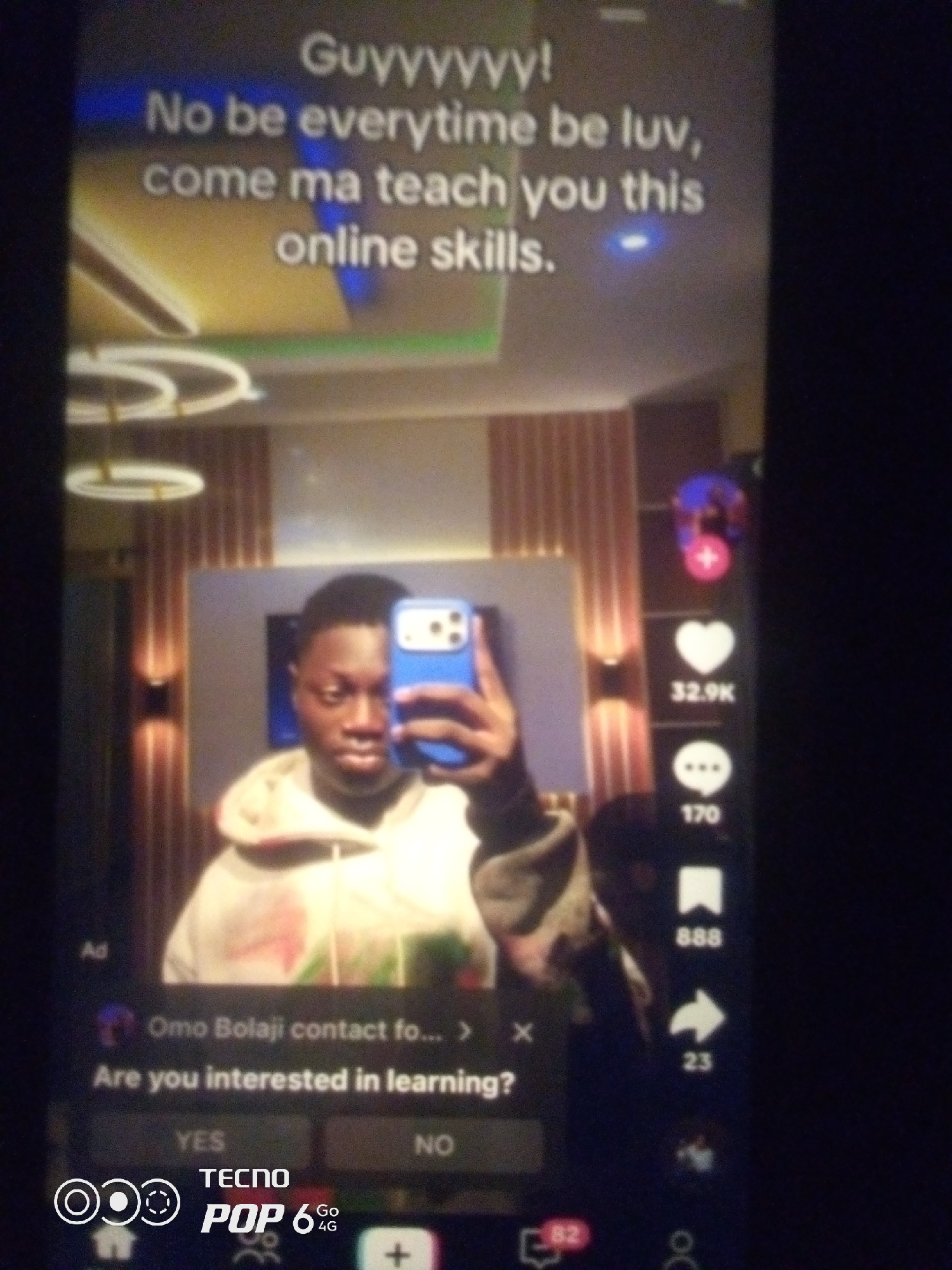 The image shows a screenshot of a mobile phone screen, likely from a social media application like TikTok. The user is taking a selfie in what appears to be a well-lit room with modern decor, possibly a home or a studio. Behind the user, there is a large screen displaying text that reads, "Guyyyy! No be everytime be luv, come ma teach you this online skills." The user is a young Black male wearing a light-colored hoodie. He is holding a blue smartphone up to take the picture. On the right side of the screen, there are social media interaction icons, including a heart with a count of "32.9K," a speech bubble with "170," a bookmark with "888," and an arrow with "23." At the bottom of the screen, there is an advertisement or a prompt asking, "Are you interested in learning?" with "YES" and "NO" options. The bottom corners of the screen display branding for "TECNO POP 6 4G." The image does not contain any information that would allow for the identification of a specific city or location within Nigeria.