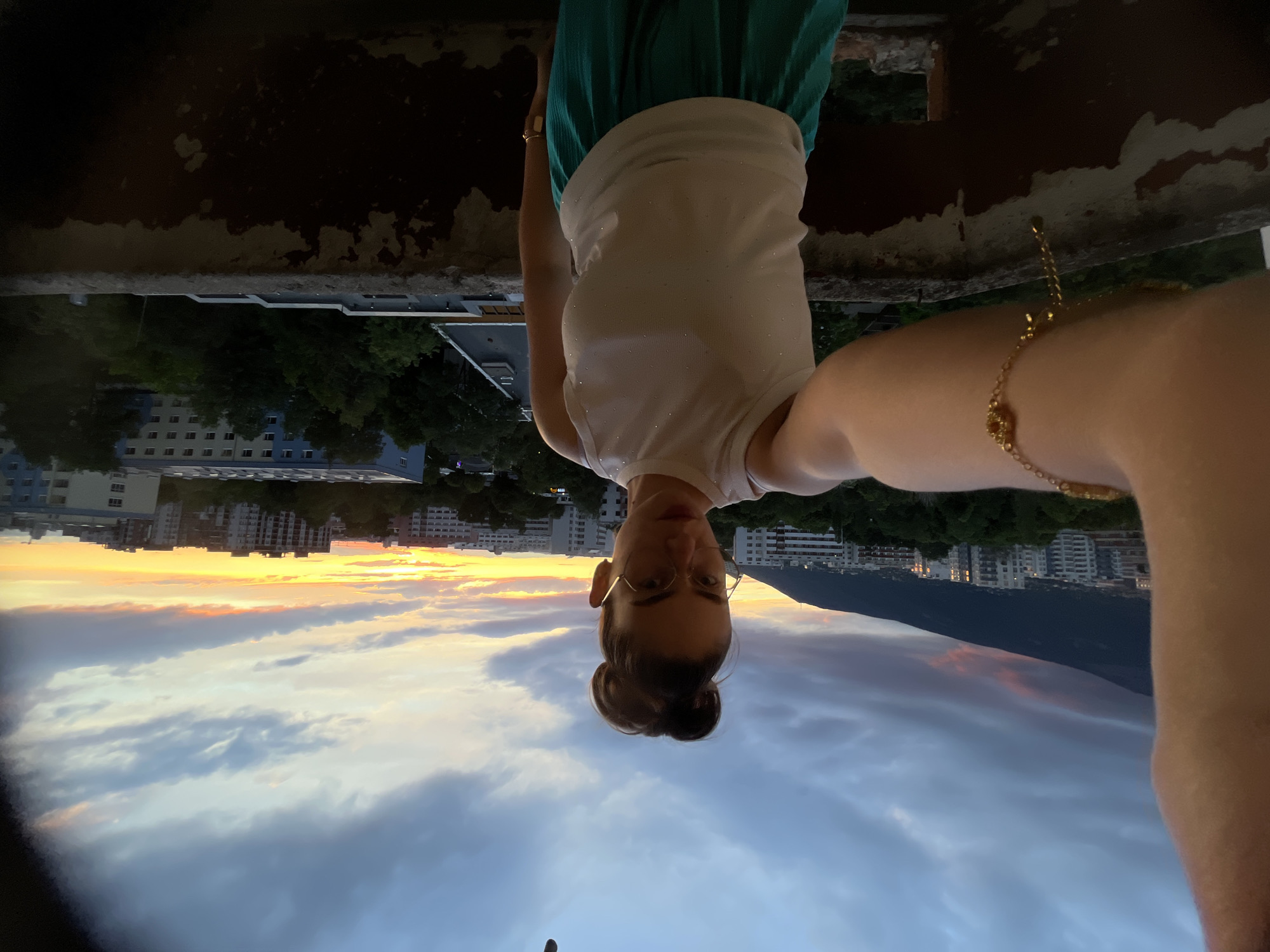 A selfie captures a young woman standing on a balcony against a backdrop of a city skyline at dusk. The woman, facing the camera with her left arm extended, wears a white sleeveless top adorned with small, glittering embellishments and teal pants. Her hair is styled in a high bun, and she sports large-rimmed glasses. She has a delicate gold bracelet on her left wrist and a similar band on her right wrist.
The balcony, constructed of weathered concrete with peeling paint, offers a vantage point over the urban landscape. Buildings of varying sizes and architectural styles populate the scene, some appearing residential, while others are larger structures. Lush green trees punctuate the cityscape, suggesting a presence of parkland or residential greenery.
Dominating the background is a majestic mountain, its dark silhouette contrasting with the vibrant hues of the sunset. The sky is a dramatic canvas of clouds, with streaks of orange, pink, and yellow illuminating the horizon, indicating the sun is either rising or setting. The overall impression is one of a serene moment captured during a transitional time of day, overlooking a sprawling metropolitan area.