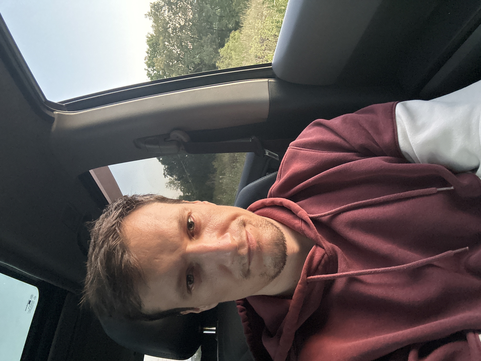 A man with short, brown hair and a goatee is seated in the passenger seat of a car, looking directly at the camera. He is wearing a maroon hooded sweatshirt with white sleeves. The car's sunroof is open, and through the open window, a field with tall grass and trees can be seen under a clear sky. The man's expression is neutral, with his eyes slightly narrowed. The interior of the car, including the dashboard and seatbelt, is visible. The lighting suggests it is daytime, possibly late afternoon. The scene appears to be in a rural or semi-rural setting, as indicated by the natural landscape outside the car. No specific details pinpoint the location to the city of Makiv, Ukraine.