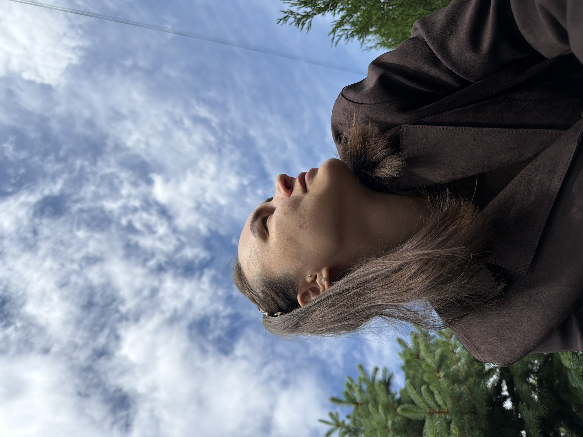 A woman in a brown coat looks up at a cloudy blue sky. Some trees are visible in the foreground. The exact location in Chaikovsky, Russia, cannot be determined from this image.
