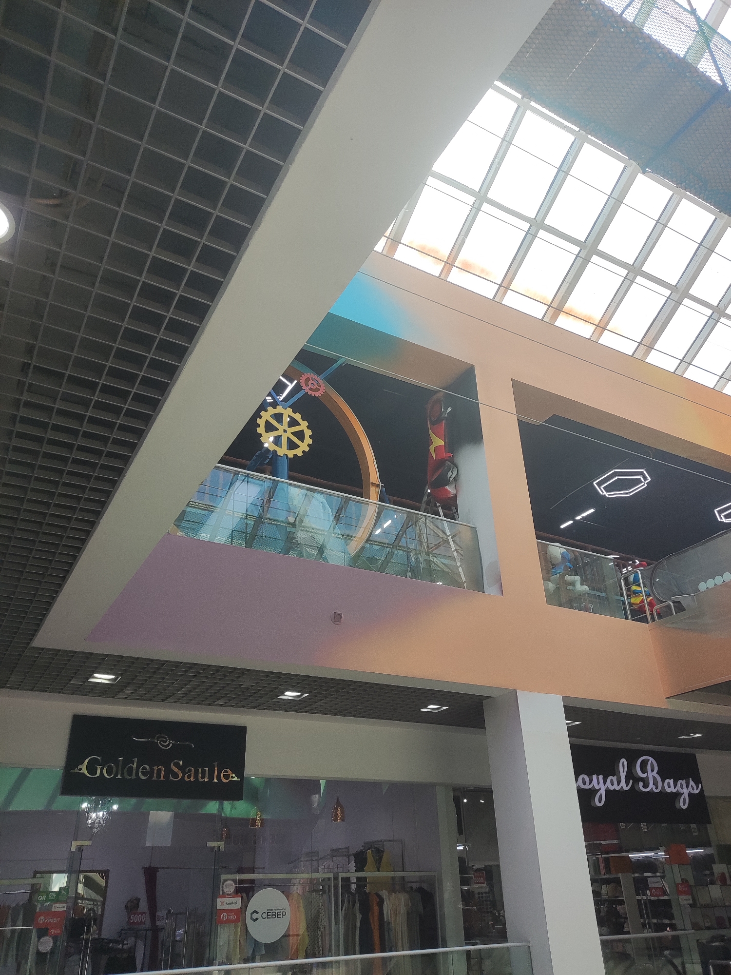 The image shows the interior of a modern shopping mall. A large, multi-story atrium is visible, with a glass ceiling that allows natural light to filter in. The atrium features decorative elements, including large yellow and red gears on a higher level, and a geometric light fixture on the ceiling of a lower level. Balconies with glass railings overlook the open space. Two shops are visible at the lower level with prominent signage: "Golden Saule" and "Royal Bags". The "Golden Saule" store appears to be a clothing boutique, with racks of garments displayed. The "Royal Bags" store is also visible, with merchandise likely related to bags and accessories. The overall environment suggests a commercial setting within a well-maintained public space. The presence of these specific store names and the architectural style are indicative of the City of Shymkent, Kazakhstan, a location known for its modern shopping centers.