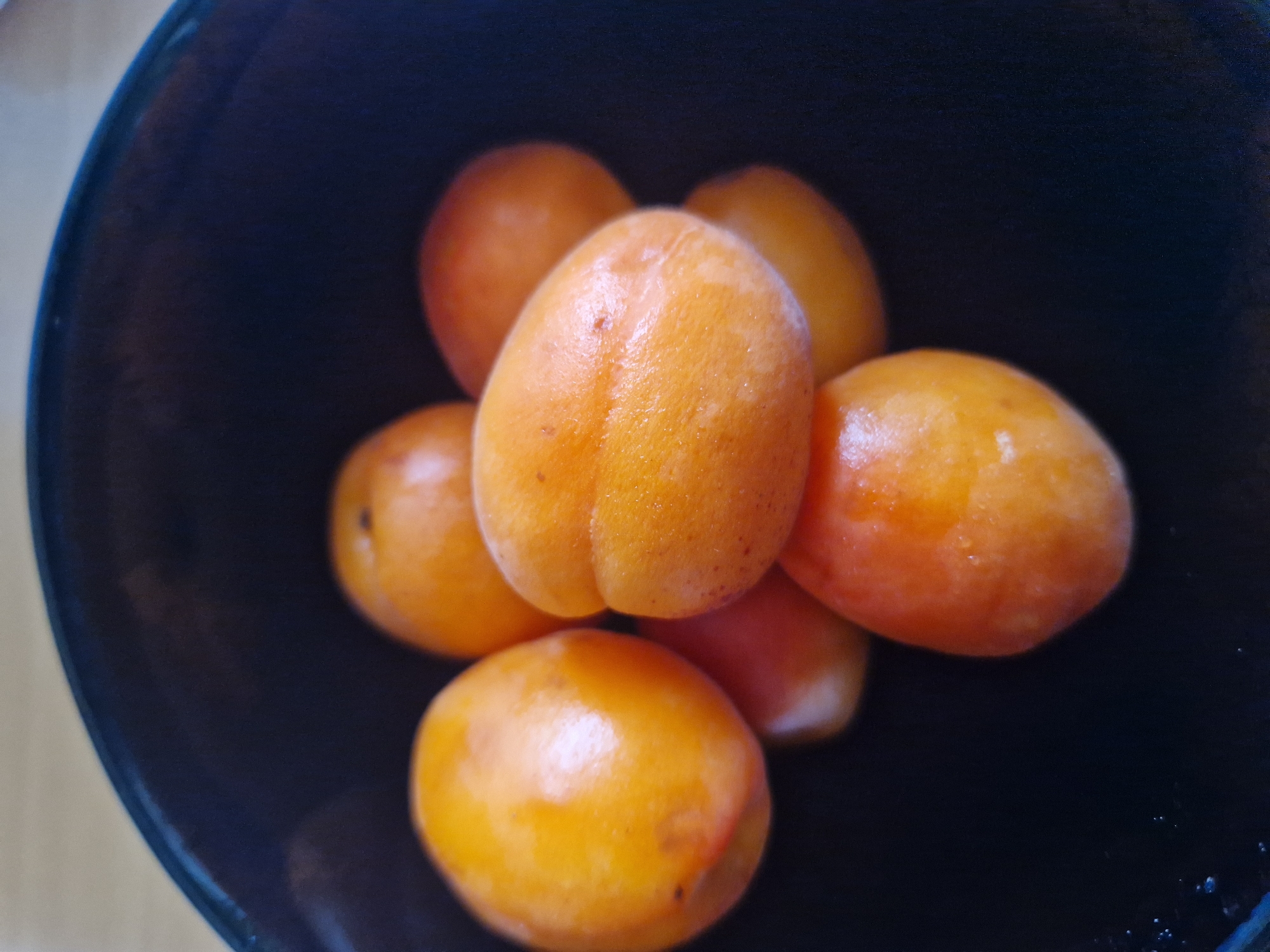 A close-up, overhead shot shows a dark bowl filled with several ripe apricots. The apricots are a vibrant orange-yellow color, with some showing a blush of red. They are plump and smooth, and appear to be freshly washed, with a slight sheen on their skins. The bowl is dark, almost black, and its rim is visible at the top of the frame. The background is out of focus, suggesting a shallow depth of field. The lighting is soft and natural, highlighting the texture and color of the fruit. The scene is simple and intimate, focusing entirely on the beauty of the apricots.