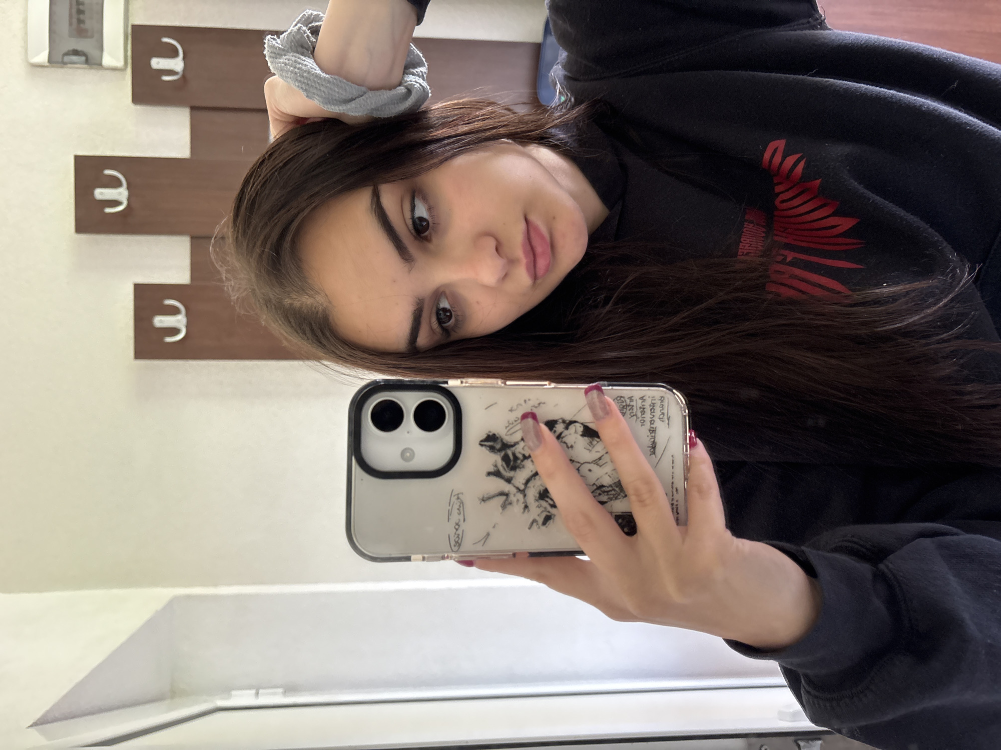 The image is a selfie taken in what appears to be an interior space, possibly a hallway or restroom, in an indoor location in the city of Polena, Bulgaria. The woman is holding a smartphone in her right hand to take the picture. The phone has a white case with a black sketch design. The woman has long brown hair and is wearing a black sweatshirt with a red graphic. A gray hair tie is on her left wrist. In the background, a wall features a series of wooden panels with metal hooks, suggestive of a coat rack. Further in the background are elements that could be a mirror or window, based on their reflective surface. The overall lighting is bright and diffuse.