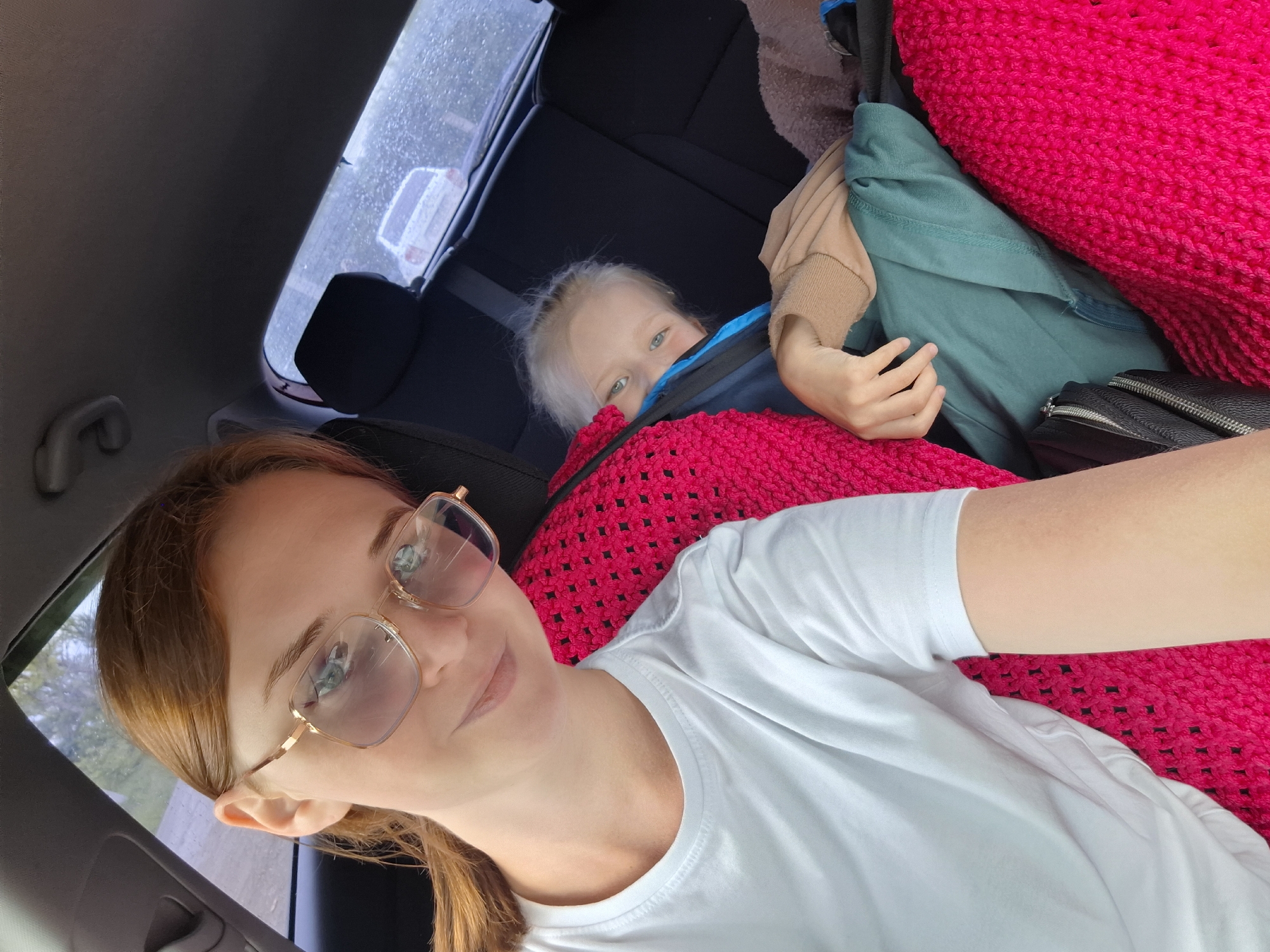 An upward-angled selfie captures a woman and a child inside a car, likely in Фоки, Russia. The woman, positioned in the foreground, has reddish-brown hair tied back, wears gold-rimmed glasses, and a plain white t-shirt. She has a subtle smile and a relaxed expression, looking towards the camera.
Behind her, partially obscured by a bright fuchsia pink, coarsely knitted blanket or garment, a young child with light blonde hair and blue eyes peeks out. The child's face shows a curious and playful expression, looking directly at the viewer. The pink knitted item covers a significant portion of the child's body and is also partially visible around the woman.
The setting is the interior of a car, with dark upholstery and headrests visible. A seatbelt is also discernible. Other items in the background include a beige/tan long-sleeved top and a teal/green hooded garment, along with a black bag featuring silver zippers. Through the car window in the upper left background, water droplets are visible on the glass, suggesting it is raining or has recently rained. A blurred view of an outdoor scene, including a light-colored vehicle, can be seen beyond the window. There is no visible text in the image. The overall mood is casual and comfortable, possibly during a car journey.