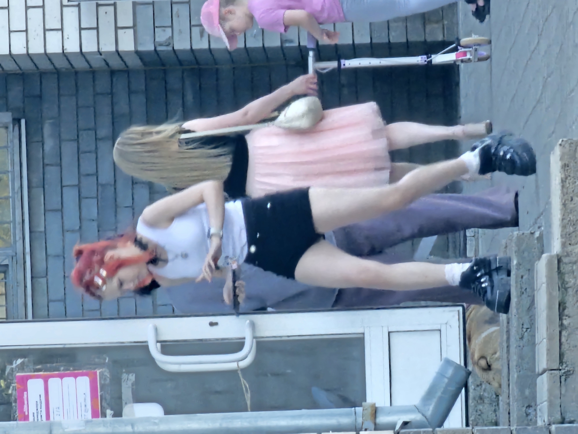 The image shows three people outdoors in what appears to be a public space, possibly an urban setting given the brick wall and paved ground. The image is rotated 90 degrees counter-clockwise, making the scene appear vertical.
In the foreground, a woman with bright red hair and bangs is leaning forward. She is wearing a white tank top, black shorts, and black shoes with white socks. She has distinctive silver hair accessories. She appears to be looking down and slightly to her right, with her mouth open.
Behind her, another person, possibly a woman, is wearing a light pink, tiered skirt and a white top. Her arms are outstretched, and she is holding a light-colored, possibly stone, object on her right arm.
Further back, a child wearing a pink hat and a pink t-shirt is visible. They are also leaning forward and appear to be interacting with the person in the pink skirt. A scooter with white and red accents is positioned between the child and the person in the pink skirt.
The setting includes a brick wall in the background and a large window or storefront in the lower portion of the image. There is also a metal railing and some concrete elements visible. The lighting suggests it is daytime.
On the left side of the storefront window, a pink and white item with text is visible. Some of the visible text includes "2/2", "T2", and what appears to be a web address or social media handle ending in "@." The overall impression is of a casual, outdoor activity, possibly involving play or a performance.