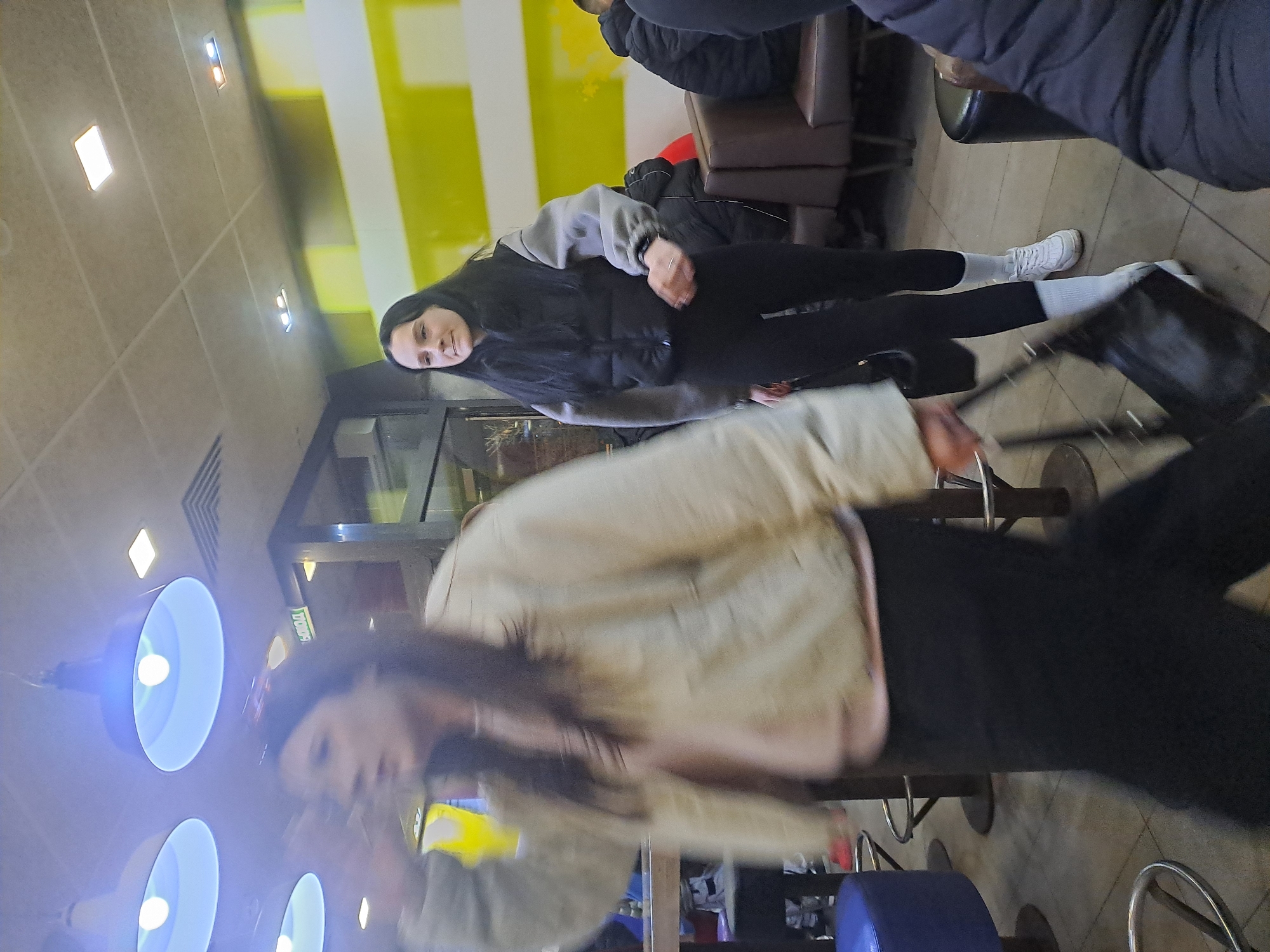 A slightly blurred, rotated indoor scene depicts several people sitting at tables. Two women are partially visible in the foreground, with one wearing a tan jacket and black pants, and the other wearing a black puffer vest with a gray hooded sweatshirt and black leggings. The second woman is wearing white shoes and socks. The background contains a yellow and white wall, with more people sitting on brown chairs. The image is oriented at an angle, possibly taken handheld and rotated. The setting appears to be in a commercial establishment, such as a fast-food restaurant. The city of Elektrostal, Russia, cannot be confirmed from this image alone.
