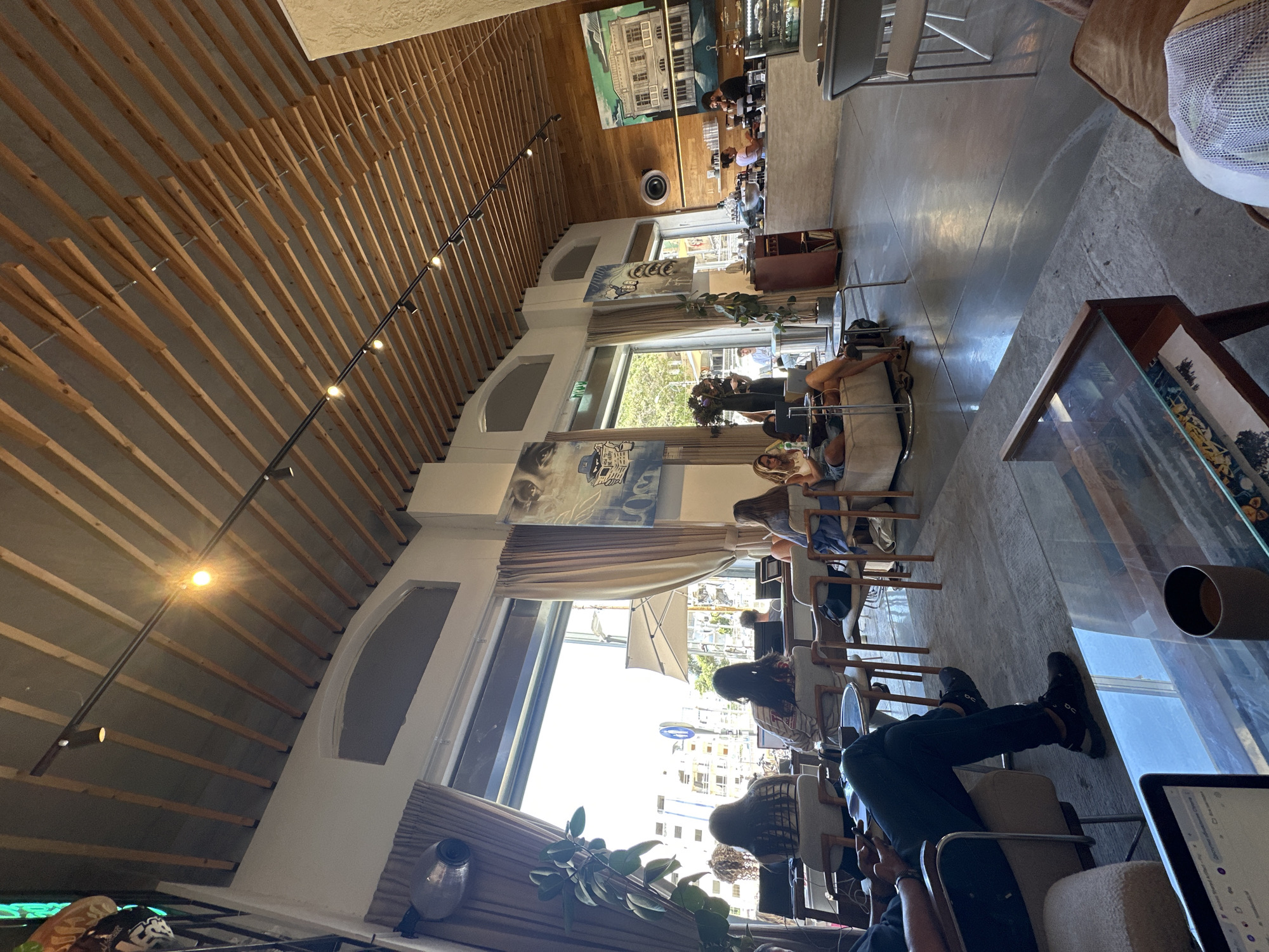 The image shows the interior of a café or restaurant in Tel Aviv, Israel. The establishment is characterized by a high, angled ceiling with exposed wooden beams and track lighting. Large arched windows allow natural light to flood the space. Patrons are seated on chairs and sofas along the windows. There is a row of people sitting with tables. At the front a person has their legs up next to a coffee table. Artwork adorns the walls.