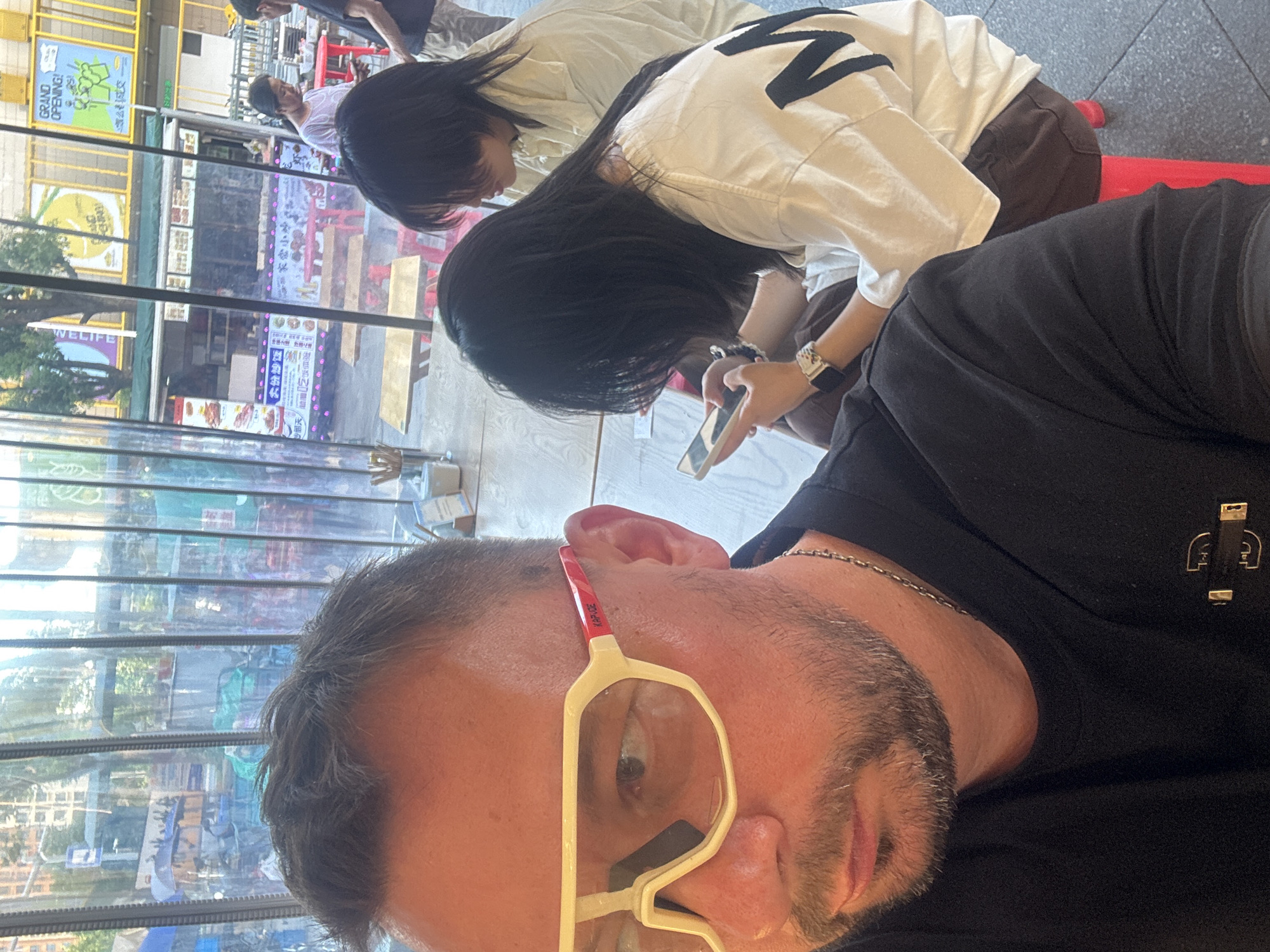 In a cafe with clear plastic partitions, a man wearing white framed glasses and a black t-shirt is taking a selfie. Behind him, two young women with dark hair are seated. One woman, in the foreground, is looking down at her phone, wearing a white t-shirt and brown shorts. The other woman is behind her, wearing a light-colored shirt. Through the partitions, a street scene is visible, with buildings, street vendors, and people. Signs in Chinese characters are present. The overall setting appears to be a bustling urban environment. The location is identified as Suzhou Industrial Park, China.