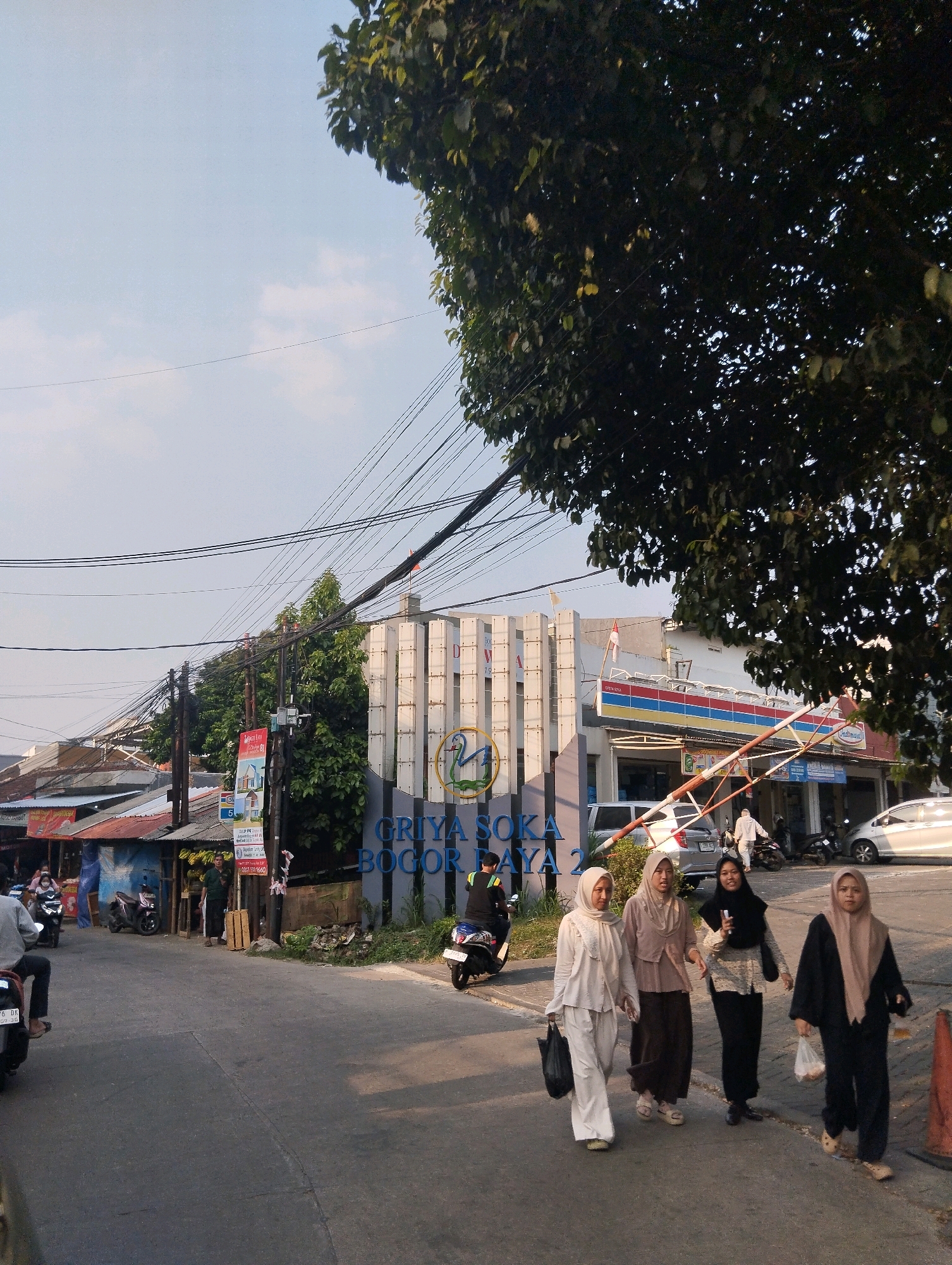 The image depicts a street scene in Sukaraja, Indonesia, with a clear sky and a large tree in the foreground. On the right side of the street, a modern building with vertical white panels serves as an entrance to "GRIYA SOКА BOGOR RAYA 2". A red and blue striped awning, likely for a convenience store, is visible above a barrier gate. Several people are walking on the sidewalk. Four women, dressed in hijabs and modest clothing, are walking towards the viewer. The woman on the far left wears a light cream outfit, the next woman a mauve outfit, the third woman a patterned top and black skirt, and the woman on the far right a black outfit with a beige hijab. Bags are visible in the hands of some of the women. To the left of the entrance building, a smaller building with a corrugated metal roof is visible, along with a street vendor and motorcycles. A man is seen riding a motorcycle on the left side of the street, and another motorcycle is parked near the vendor. The street itself is paved, with some debris on the side. Power lines crisscross the sky above the scene. The overall impression is a bustling urban environment.