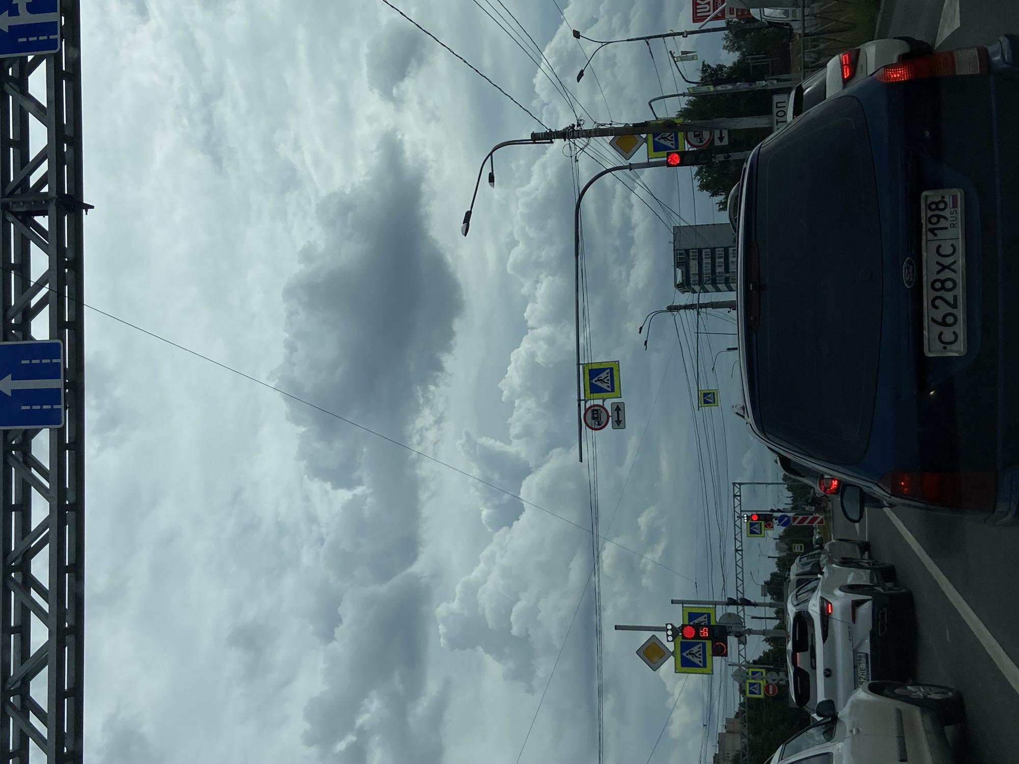 The image shows a view from inside a vehicle in traffic on a cloudy day in Saint Petersburg, Russia. Ahead, there are several cars, traffic lights, and street signs. The traffic lights are red, indicating a stop. The sky is filled with dramatic, dark clouds. A large metal overhead structure with traffic signs is visible at the top of the image. The road appears to be an urban street with buildings and trees visible in the background. The license plate of the car in front is visible and reads "c628xc 198".