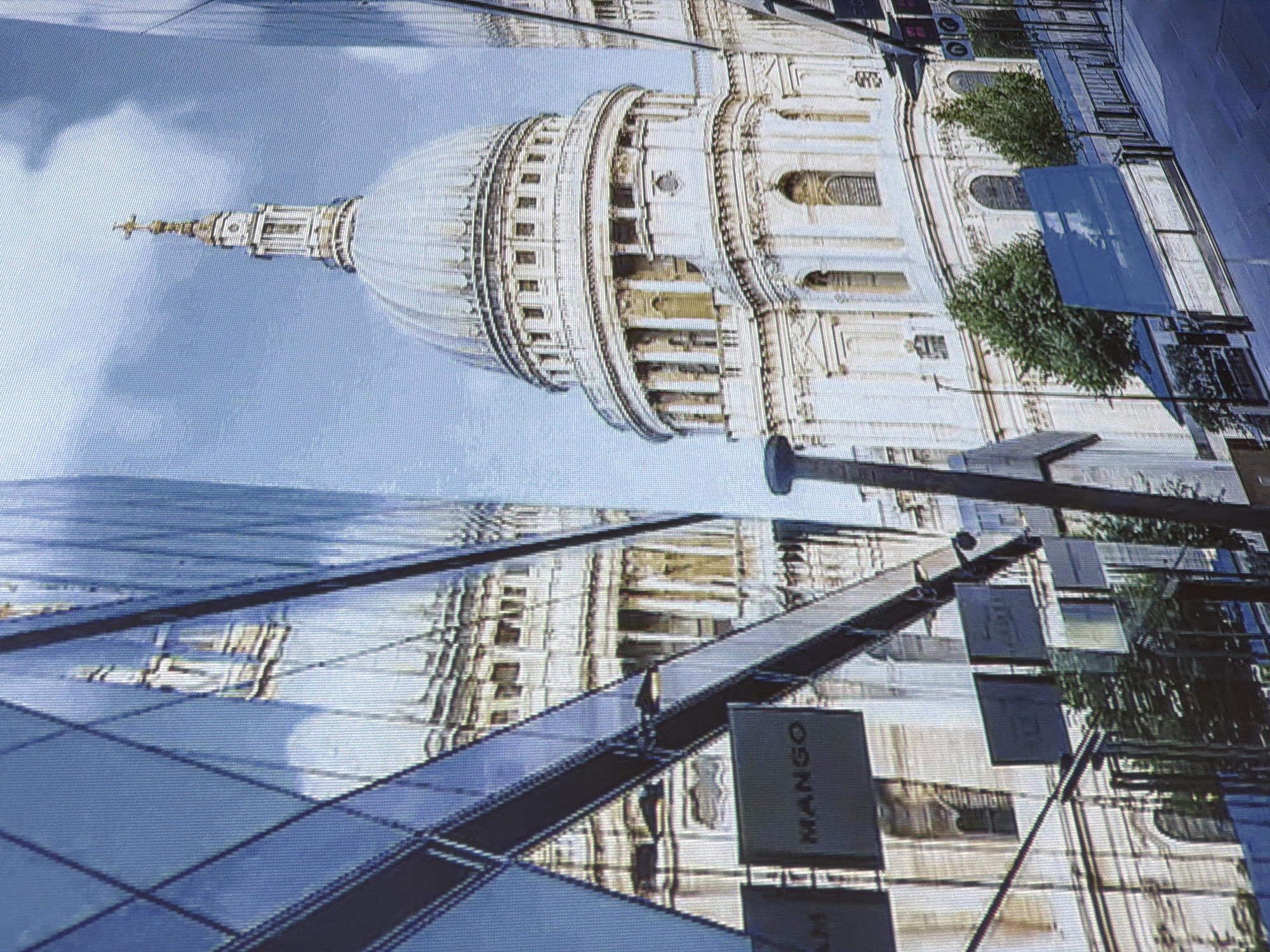 The image shows a reflection of St. Paul's Cathedral in London, England, on the glass facade of a modern building. The cathedral's dome and cross are clearly visible in the reflection. The modern building has a geometric pattern of glass panels. In the foreground, there are signs on the modern building, including one that says "MANGO". Trees and a railing are also visible. The sky is cloudy. The image is not located in Sacramento, United States of America.