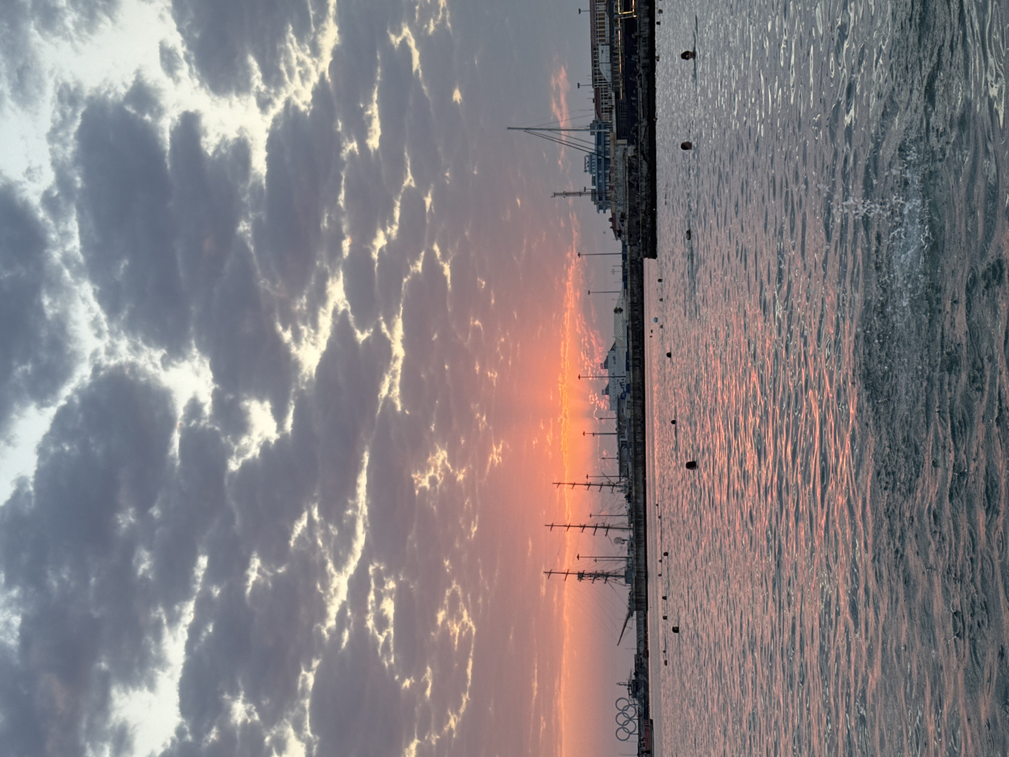 The image captures a sunset over the Black Sea in Sochi, Russia. The sky is filled with dramatic clouds, tinged with orange and pink from the setting sun. In the distance, a harbor is visible, featuring several tall-masted sailing ships, a cruise ship, and various port structures. The Olympic rings symbol is also present on the left side of the harbor. The water in the foreground is calm, with ripples reflecting the colors of the sky. Several people are swimming or wading in the water, indicated by their heads appearing above the surface. Some are closer to the shore, while others are further out. There are also buoys scattered in the water, marking a swimming area. The overall scene depicts a serene evening by the sea, with a blend of maritime activity and recreational swimming.