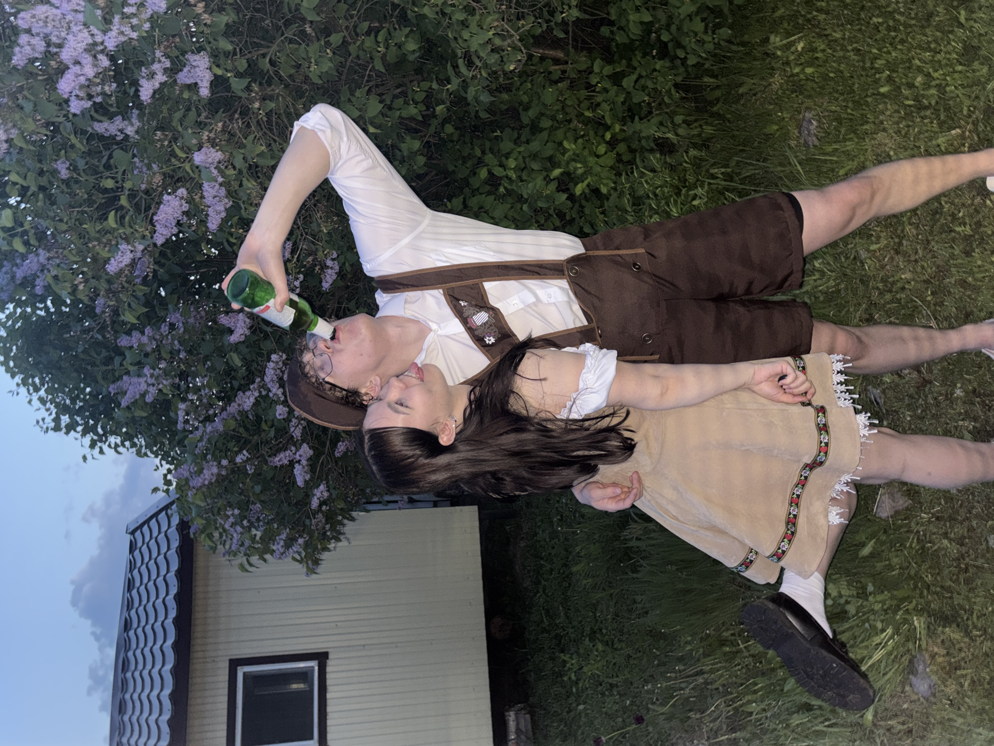 Two people, a male and a female, are lying on grass, presumably in Пальники, Russia. The male is wearing a traditional Bavarian outfit with brown lederhosen, a white shirt, and suspenders, and is drinking from a green bottle. The female is wearing a short beige dress with embroidered trim and white socks with black shoes. In the background is a lilac bush and a house with a brown roof and yellow siding. The lighting suggests it is dusk or evening.