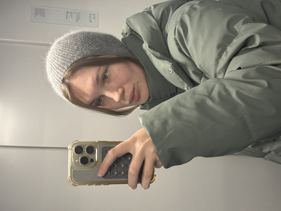 The image depicts a young person taking a mirror selfie. The photo is oriented sideways, with the subject's head towards the left and her body extending towards the right.
The central figure is a young person, likely female, with fair skin and light-colored eyes. Her light brown hair, with visible bangs, frames her face and peeks out from under a gray knit beanie. The beanie has a soft, slightly fuzzy texture, suggesting a material like angora or cashmere. She is wearing a thick, puffy winter jacket in a muted sage green or light olive color. The jacket features silver-colored snap buttons along the front placket. Her expression is calm and neutral, with a slight hint of a smile or smirk, and she looks directly at the phone's camera lens.
Her left hand holds a smartphone, extended towards the mirror to capture the reflection. The smartphone appears to be a model with a prominent triple-lens camera array on the back, encased in a clear or translucent protective case with metallic gold edges. A circular pop socket or similar grip accessory with multiple small bumps is attached to the back of the phone case.
The background is a plain, light-colored surface, indicative of an indoor setting, likely a room with white or very light gray walls. The reflective surface of the mirror is evident. In the upper left portion of the image (which would be the upper right if the image were correctly oriented), there is a rectangular white label or sign affixed to the wall. Some faint, mirrored, and upside-down text is visible on this label, including what appears to be numbers like "09" and "20," but the full text is not legible.
The lighting is bright and even, illuminating the subject's face clearly. The overall impression is casual and contemporary, capturing a moment of self-portrayal.