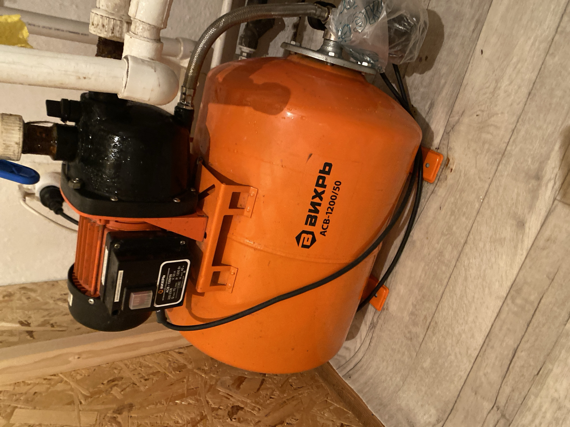 A bright orange water pump with a black motor sits on a light brown wooden floor. The pump has a cylindrical tank and a prominent black pump head. A silver flexible hose connects to the pump. The pump has a label with Russian text, including the brand name "ВИХРЬ" and the model number "АСВ-1200/50". The pump is located indoors, with a wall constructed of light brown particle board visible in the background. Pipes and electrical components are also visible near the pump. The scene is well-lit, suggesting it might be a utility room or basement.