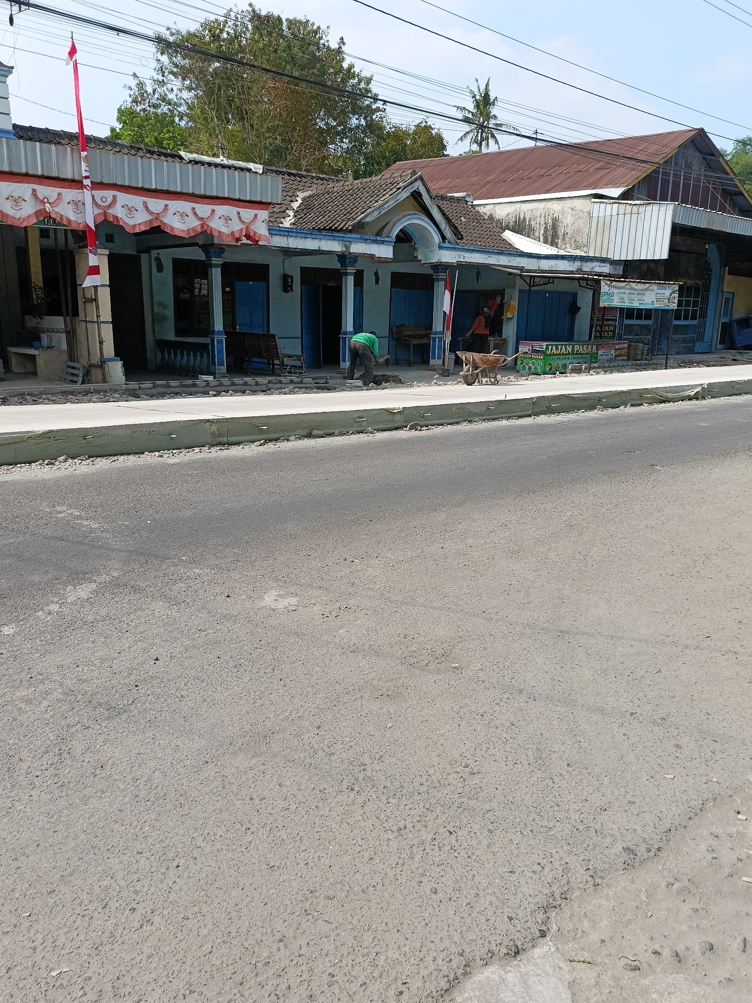 The image shows a street scene in Barukan, Indonesia, with buildings on the left and the road in the foreground. The buildings appear to be small shops or homes with blue doors and windows and decorative awnings. There is a red and white flag hanging on the left side of one of the buildings. A person in a green shirt is seen working on the sidewalk in front of the buildings, and a wheelbarrow is nearby. The road is paved with asphalt and shows signs of wear. Power lines are visible crisscrossing the sky above the buildings. The overall impression is one of a developing area with ongoing construction or maintenance work.