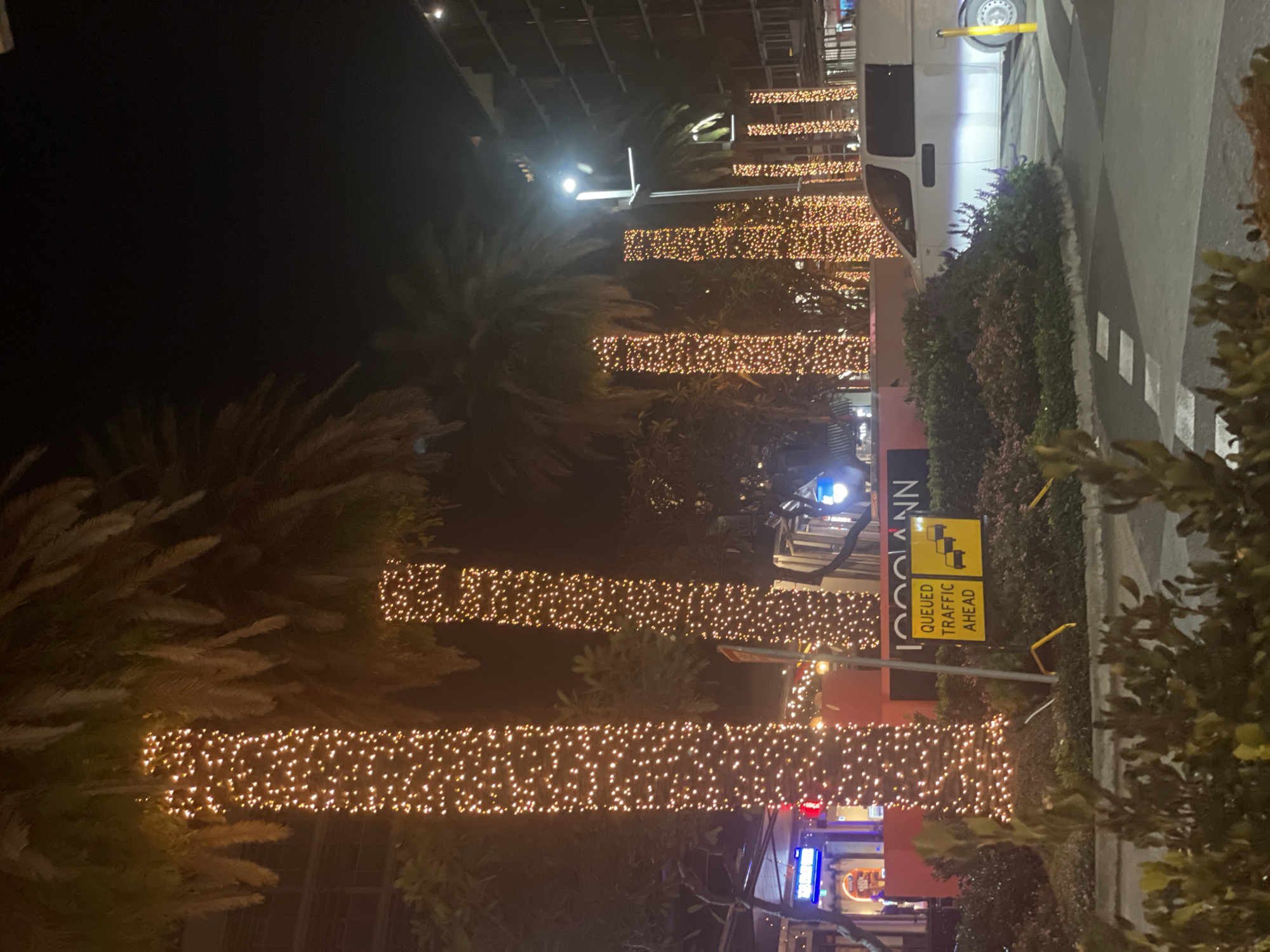 The image depicts a nighttime scene in Brisbane, Australia, likely in a commercial or entertainment district, given the building facades and illuminated palm trees. Several palm trees are wrapped in strings of warm-toned lights, creating a festive atmosphere. In the foreground, a white van is parked on the right side of the frame. A yellow sign with black text and graphics indicating "QUEUED TRAFFIC AHEAD" with three car icons is visible on a pole in the mid-ground. The background features buildings with illuminated windows, suggesting a populated area. The overall impression is one of a vibrant urban environment at night, enhanced by decorative lighting.