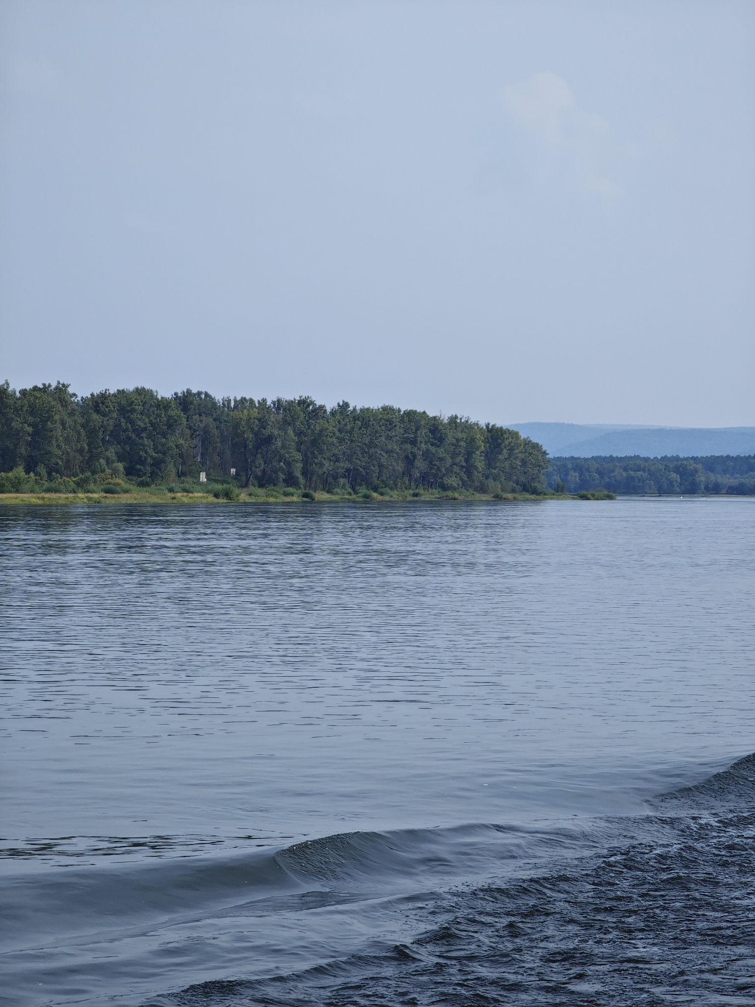 The image depicts a wide, calm river with a dense, green forest lining the distant bank. The sky is a pale blue with a few wispy clouds. The foreground of the river shows gentle ripples and a prominent wake on the right side, indicating movement, possibly from a boat. The forest on the far bank is thick with deciduous trees, and a few structures, appearing to be white signs or markers, are visible amongst the trees. Beyond the initial treeline, the landscape recedes into a hazy, blue-tinted vista of rolling hills. The overall scene is serene and natural, characteristic of a riverside landscape in a rural or semi-rural setting. The location is identified as Kononovo, Russia. There are no people or animals visible in the image.