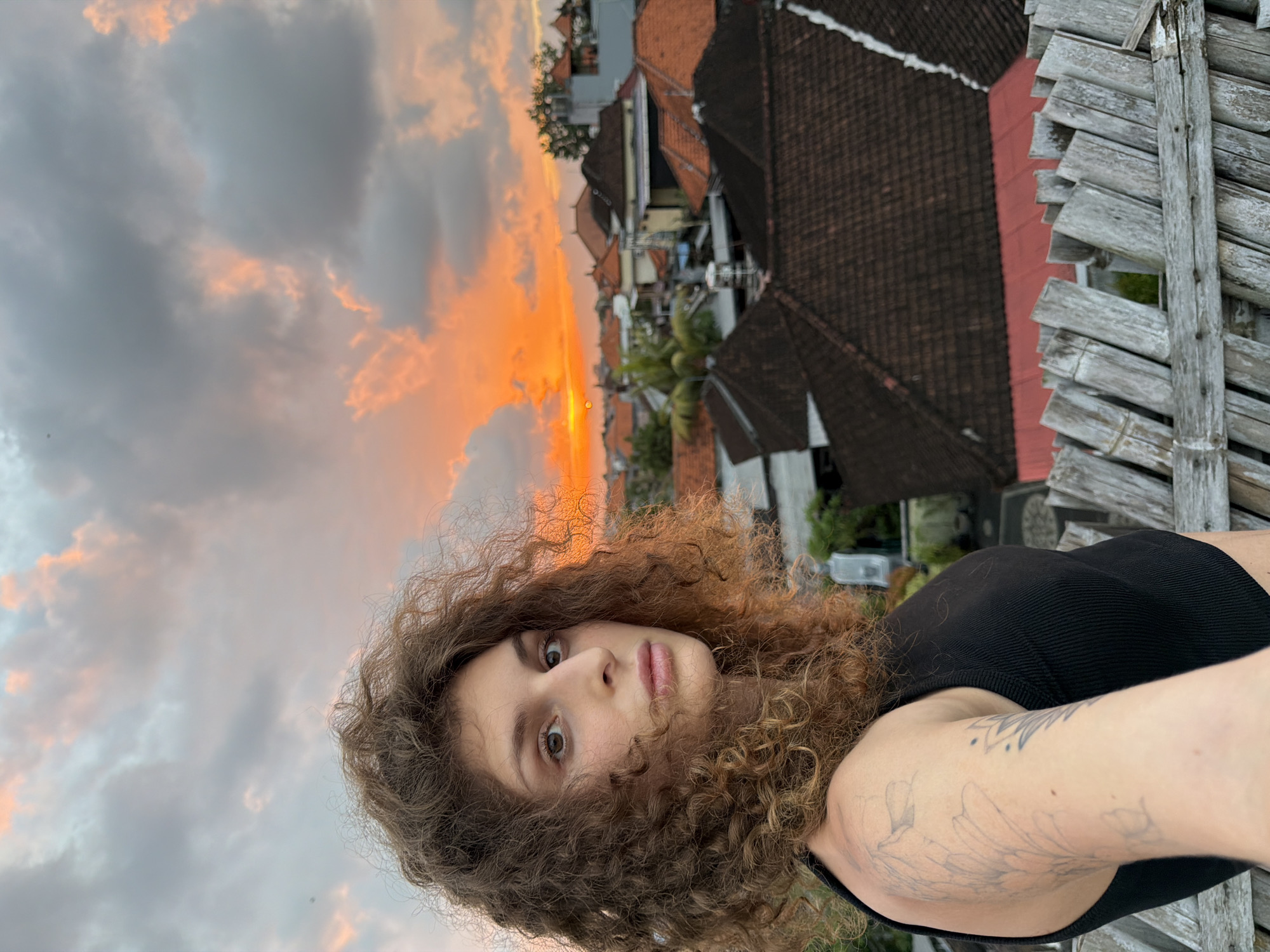 This is a portrait of a young woman with curly, auburn hair. She is looking directly at the camera with a neutral expression. She is wearing a black tank top. Her left arm is visible, and she has a tattoo of flowers on it. The background suggests an outdoor setting, possibly a rooftop, with traditional Indonesian architecture, including terracotta roofs. The sky is dramatic with shades of orange and grey clouds, indicating sunset or sunrise. The image appears to be a selfie taken with a phone. The time of day is likely sunset. The location is suggested by the architecture to be North Kuta, Indonesia.
