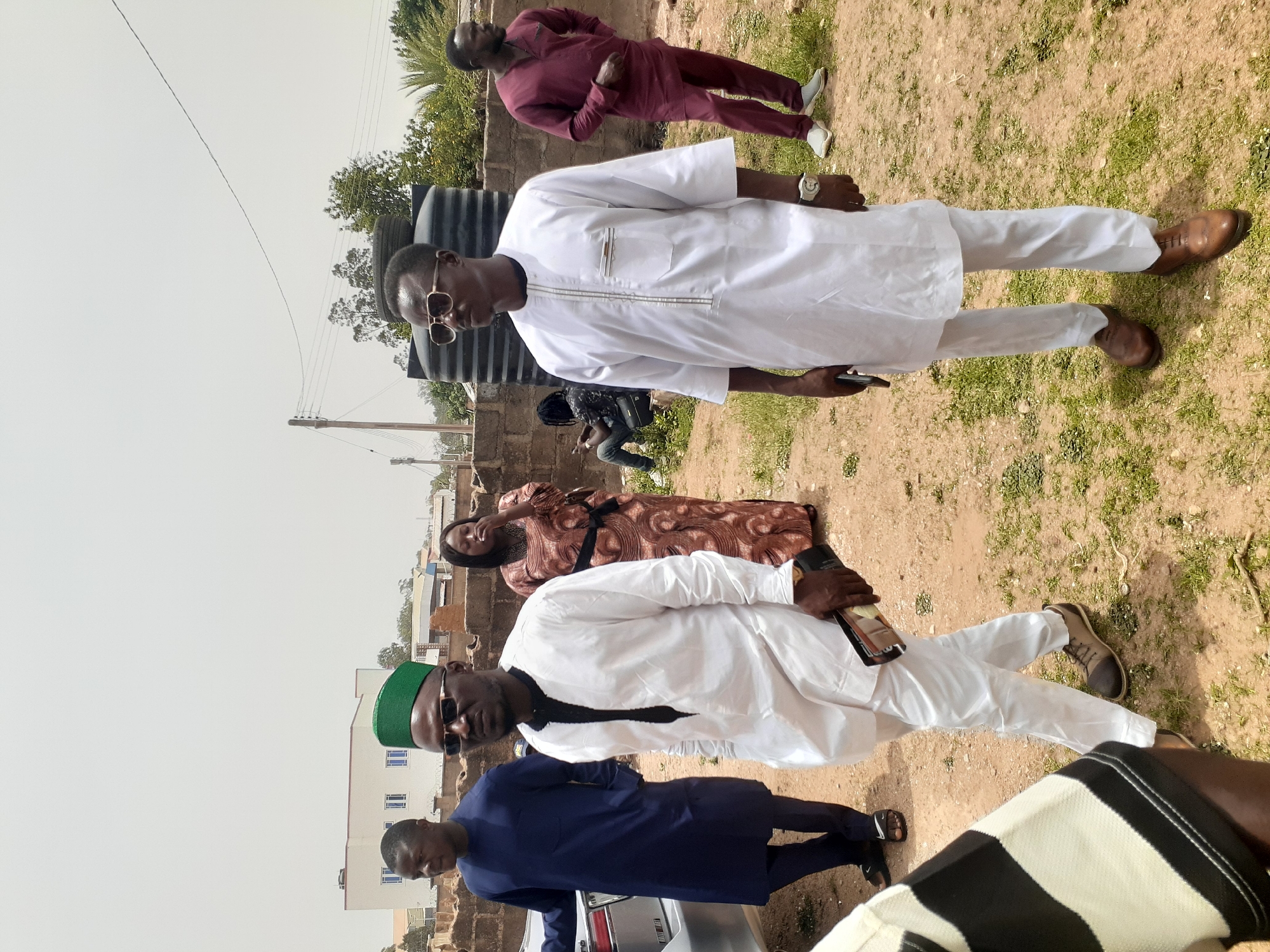 The image shows a group of people outdoors in what appears to be a residential area of Barakin Lerawi, Nigeria. The ground is dry with patches of grass.
Several individuals are present. Two men stand prominently, wearing white traditional attire. One man wears a green cap. Another man in a maroon outfit stands in the background. A woman in a patterned brown dress sits in the background. Another man is wearing a blue outfit.
Other elements include a water tank, a brick wall, electrical poles and wires, and a partial view of buildings in the distance. A silver car is also visible in the image.