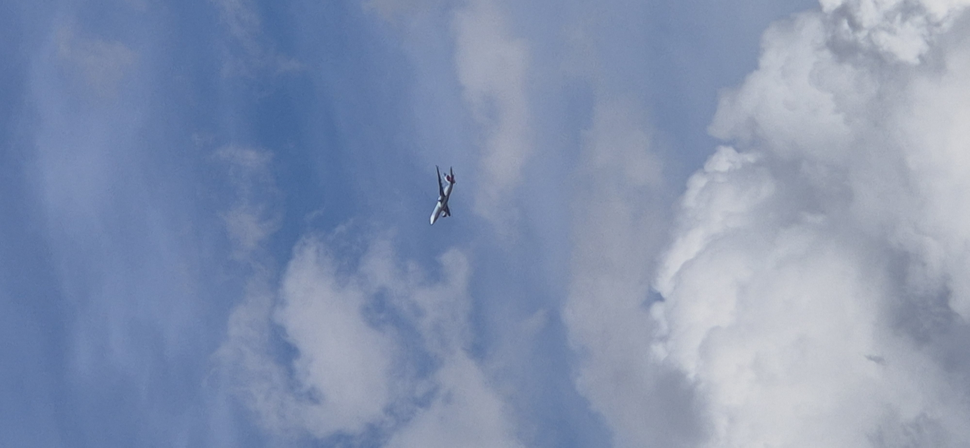 An airplane is visible in the sky above Mainz, Germany. The airplane is white with red accents on its tail and appears to be in flight. The sky is blue with scattered white clouds. The image is taken from below, looking up at the airplane. The clouds are a mix of fluffy cumulus and wispy cirrus formations. The overall scene is one of a clear day with an airplane passing overhead.