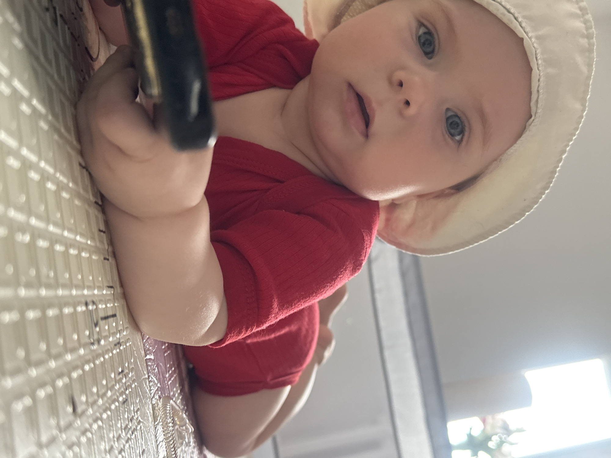 A baby wearing a red shirt and white hat is lying on a patterned surface. The baby is holding a black object in its left hand. A window is visible in the background. The location is unspecified but Rusalevka, Russia is noted as the city.