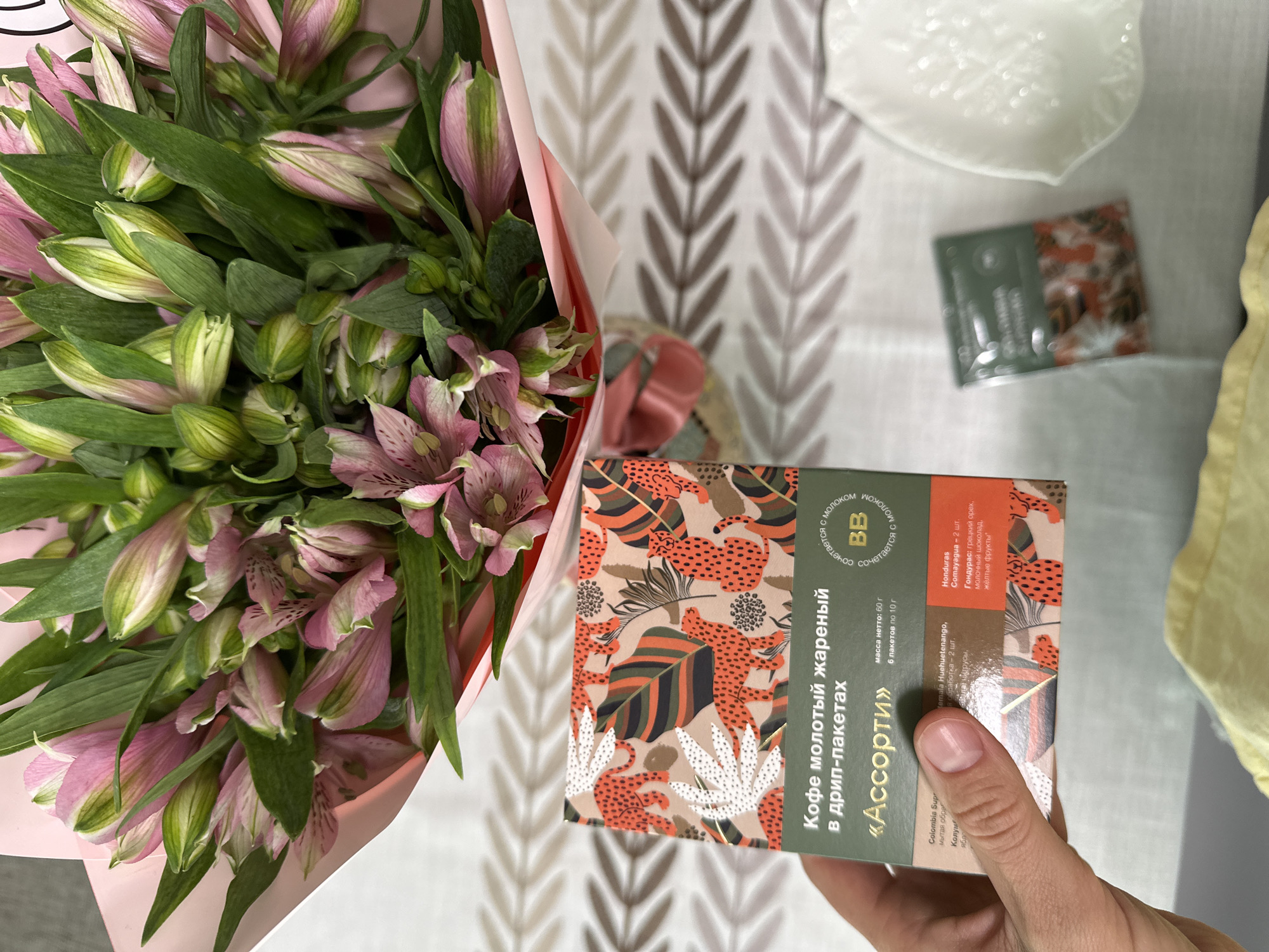 A hand is holding a box of coffee. The box is green and decorated with an animal print of leopards and tropical leaves in shades of orange, black, and green. Text on the box is in Russian and says "Кофе молотый жареный в дрип-пакетах «Ассорти»" which translates to "Ground roasted coffee in drip bags 'Assorti'". The box contains 6 drip bags of 10 grams each, for a total net weight of 60 grams. The text also mentions different coffee origins like Colombia, Guatemala Huehuetenango, and Honduras Comayagua, with flavor notes described as walnut, citrus, milk chocolate, and yellow fruits. To the right of the box, there is a smaller green packet of coffee, and further to the right, a white decorative plate. In the background, a bouquet of pink and green flowers, possibly alstroemeria, is arranged and wrapped in pink paper. The entire scene is laid out on a table with a patterned tablecloth. The image appears to have been taken indoors, likely in a home setting. The image does not contain information to determine its location in Vsevolozhsk, Russia.