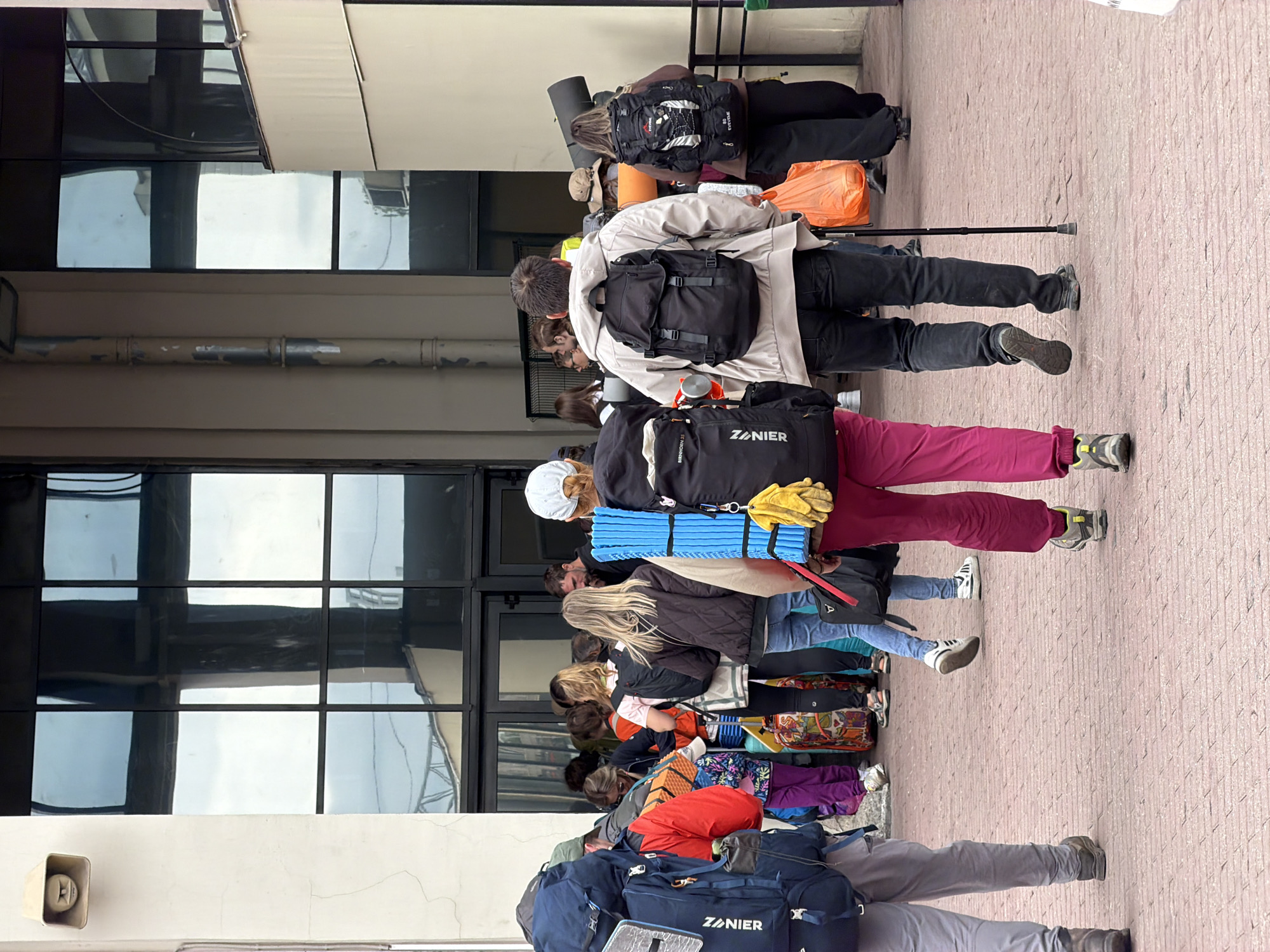 This image captures a group of people walking along a red brick pathway in front of a building with large, reflective windows. The individuals are dressed casually and many are carrying large backpacks, suggesting they are travelers or hikers. Some are also holding camping mats and bags, further indicating outdoor activity. In the background, the windows reflect the sky and surrounding structures. The overall impression is a scene of people preparing for or returning from an excursion. The building appears to be a public space, possibly a transportation hub or a venue.
The image is located in the city of Saint Petersburg, Russia. The architectural style of the building, particularly the large glass panels and the overall structure, is consistent with modern construction found in the city. The paved pathway and the presence of a group with travel gear suggest an area that experiences foot traffic and potentially serves as a departure or arrival point for trips. The weather appears to be overcast, as indicated by the reflections in the windows.